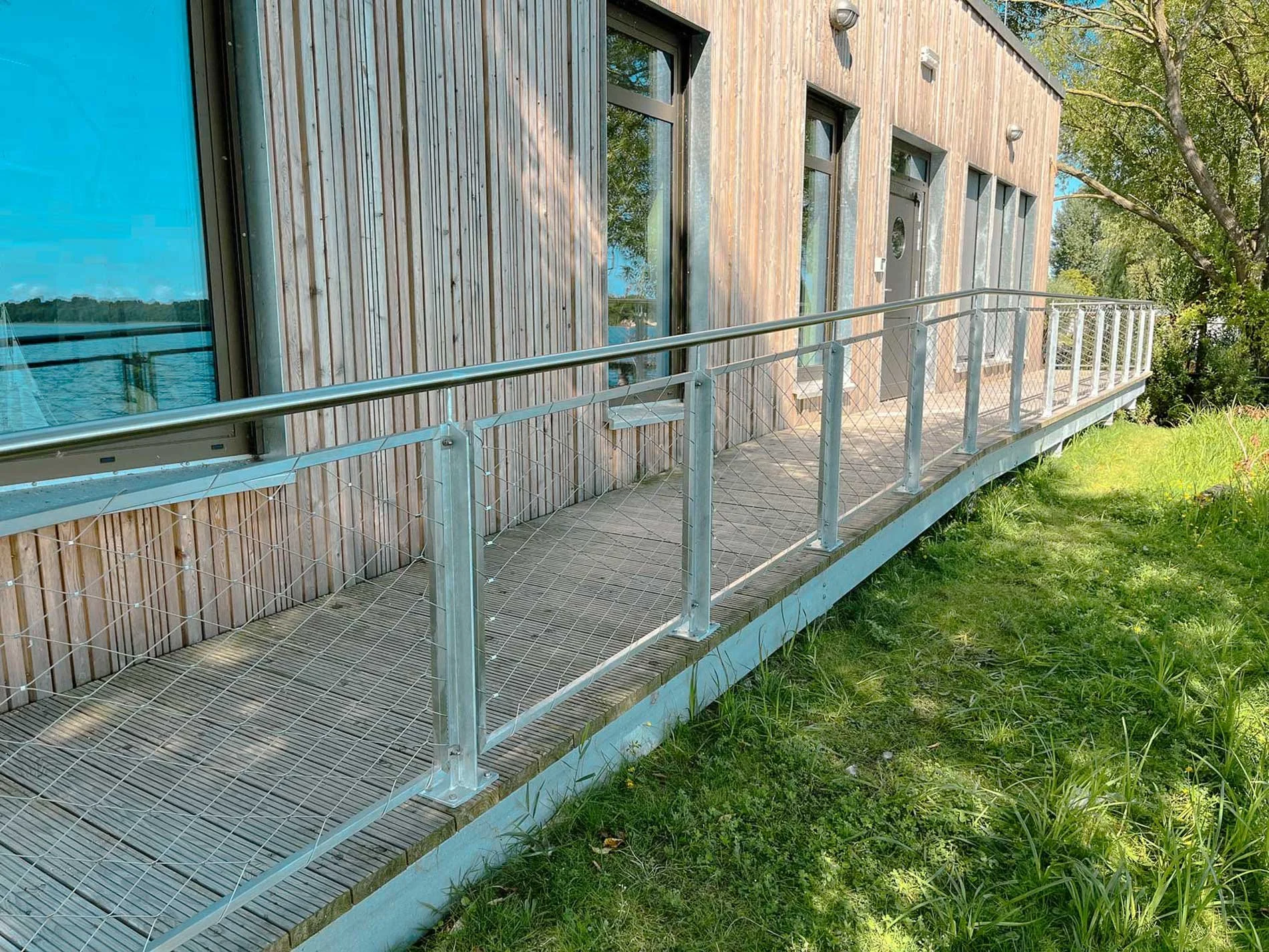 Balkon an einem modernen Gebäude mit Holzverkleidung, Geländer und mehreren Fenstern, umgeben von Bäumen und Grünfläche im Freien.