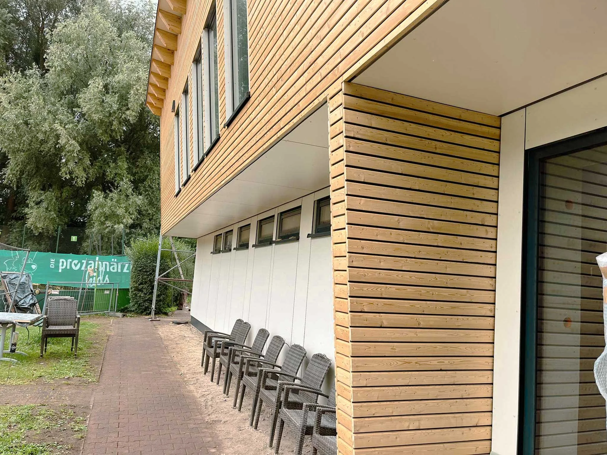Außenansicht eines modernen Gebäudes mit Holzverkleidung und großen Fenstern, umgeben von Bäumen und Gartenmöbeln.