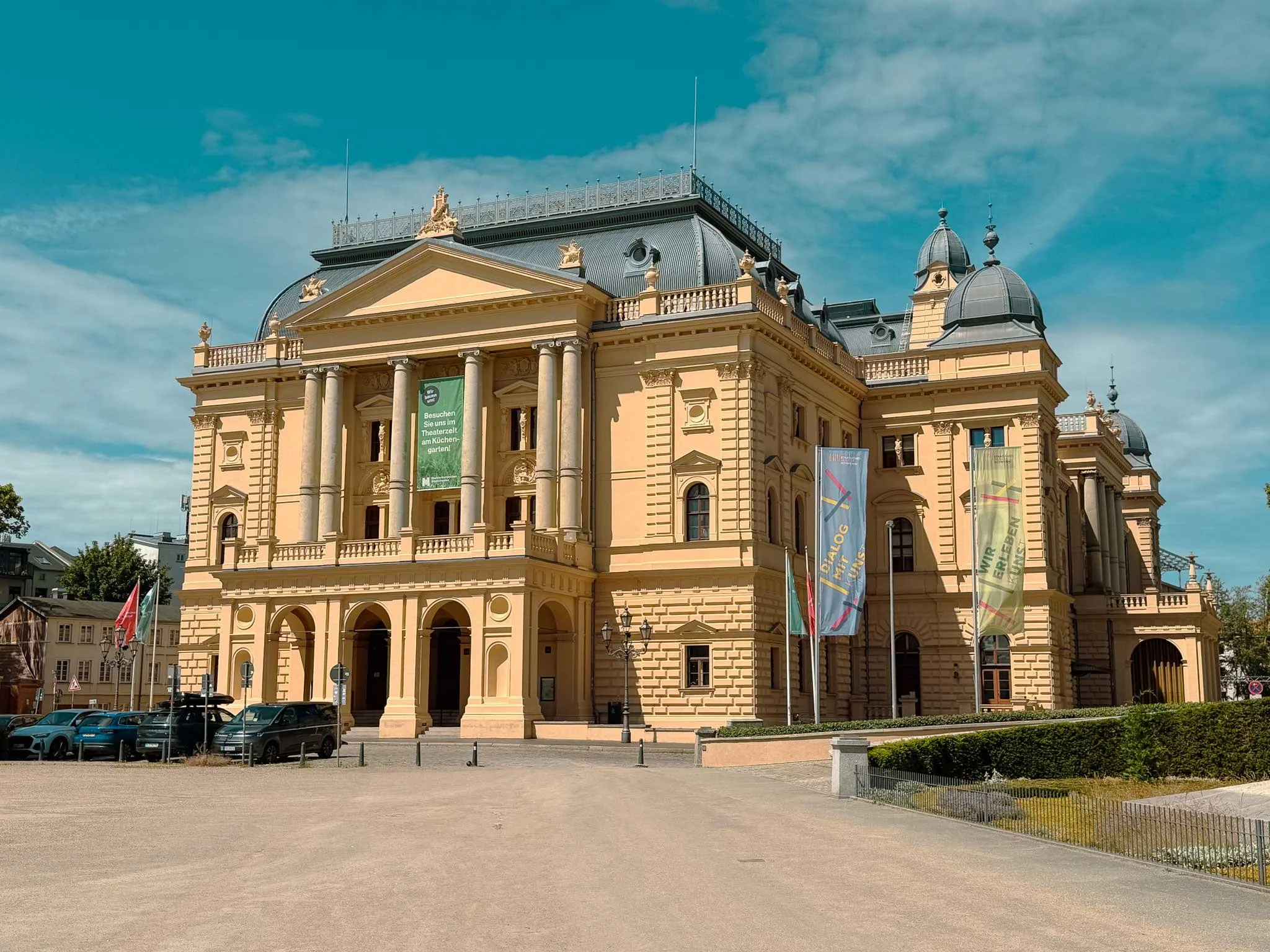 Ein barockes Gebäude mit Säulen und kunstvollen Verzierungen, mehrere Flaggen vor dem Eingang, blaue Himmel mit wenigen Wolken.