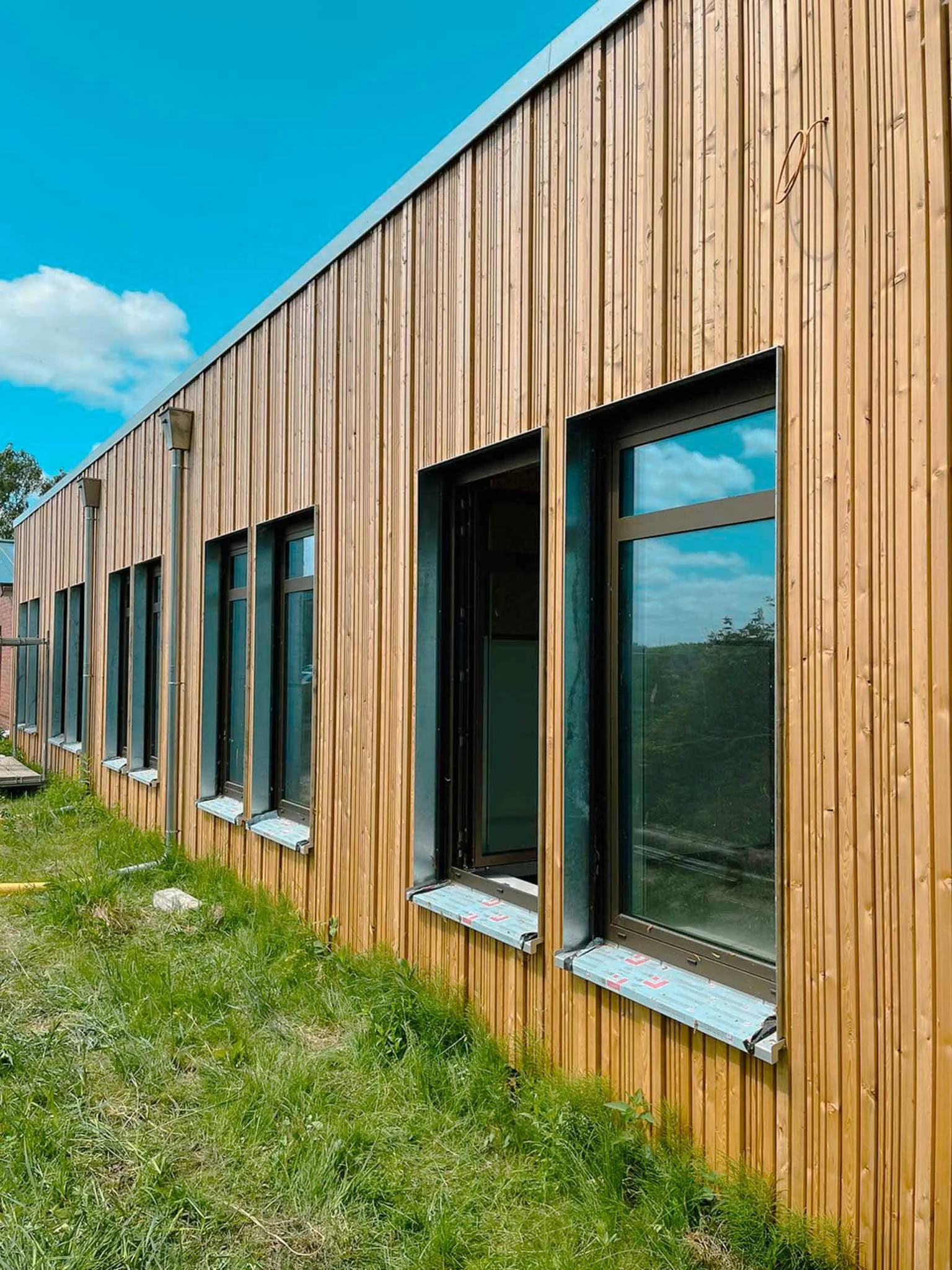 Holzverkleidetes Gebäude mit großen Fenstern und Balkonen auf einer grünen Wiese bei blauem Himmel.