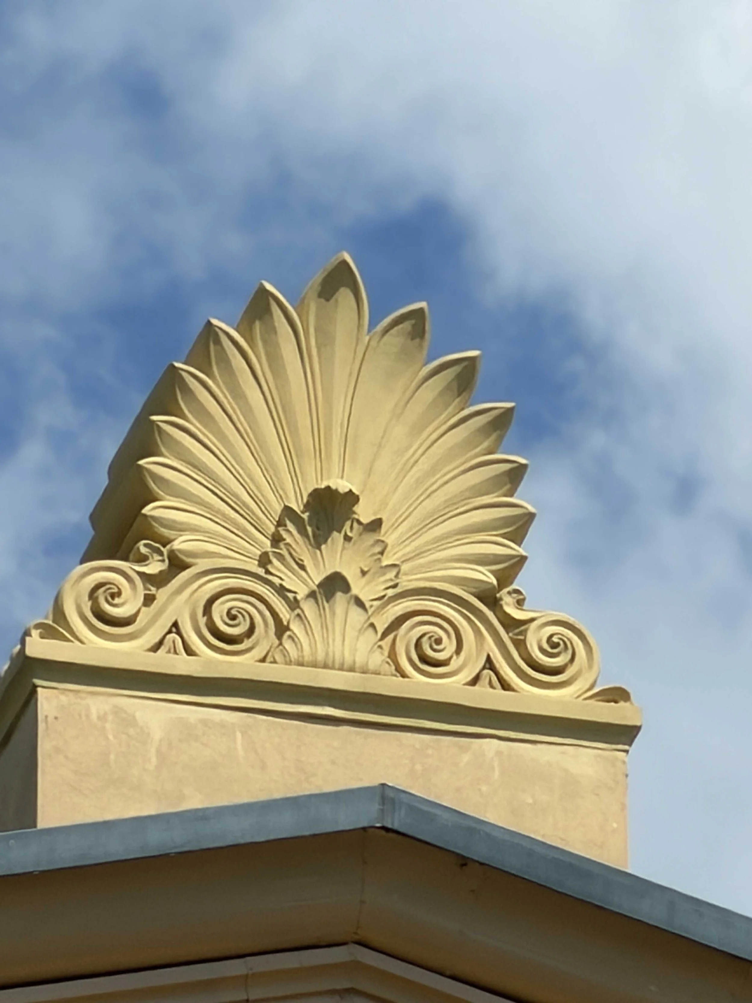 Nahaufnahme eines kunstvollen, goldfarbenen Dekorelements am oberen Teil eines Gebäudes, mit blauen Himmel im Hintergrund.