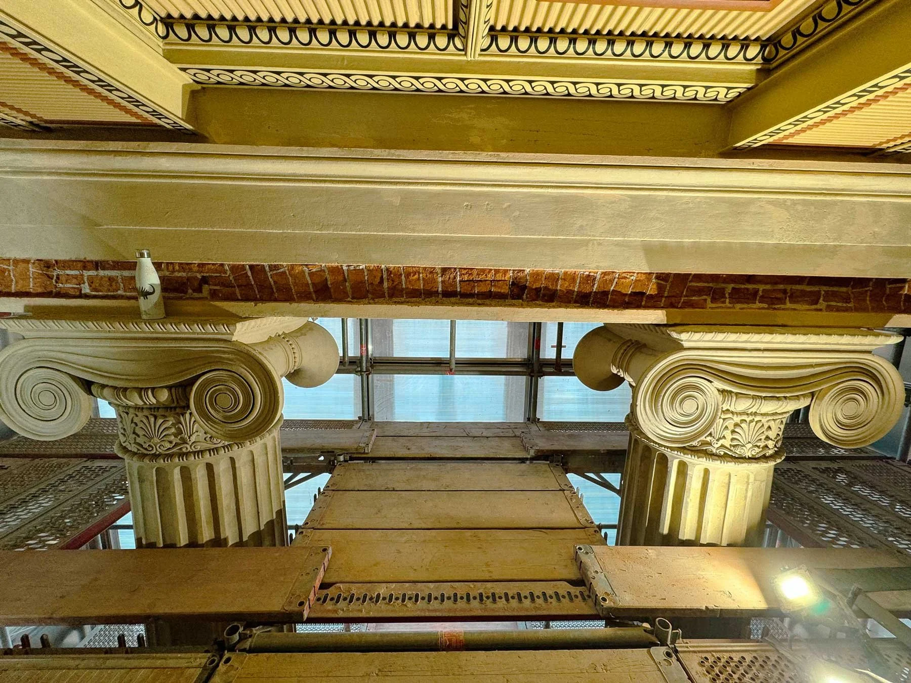 Das Bild zeigt den Blick nach oben auf zwei antike Säulen mit ionischen Kapitellen, umgeben von Baugerüsten und Schutznetzen. Im Hintergrund sind große Fenster sichtbar.