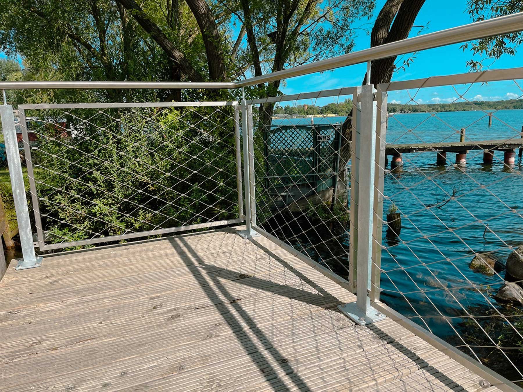 Ein Holzsteg mit einer Metallgeländer am Wasser, umgeben von Bäumen und einer blauen Himmel mit Wolken.