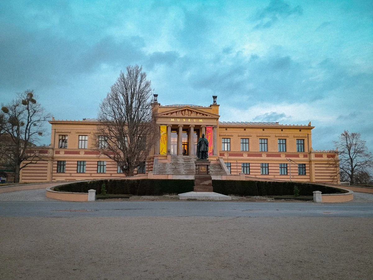 Ein historisches Museum Gebäude mit Stufen, einer Statue vor dem Eingang und Bäumen auf beiden Seiten, bei bewölktem Himmel.