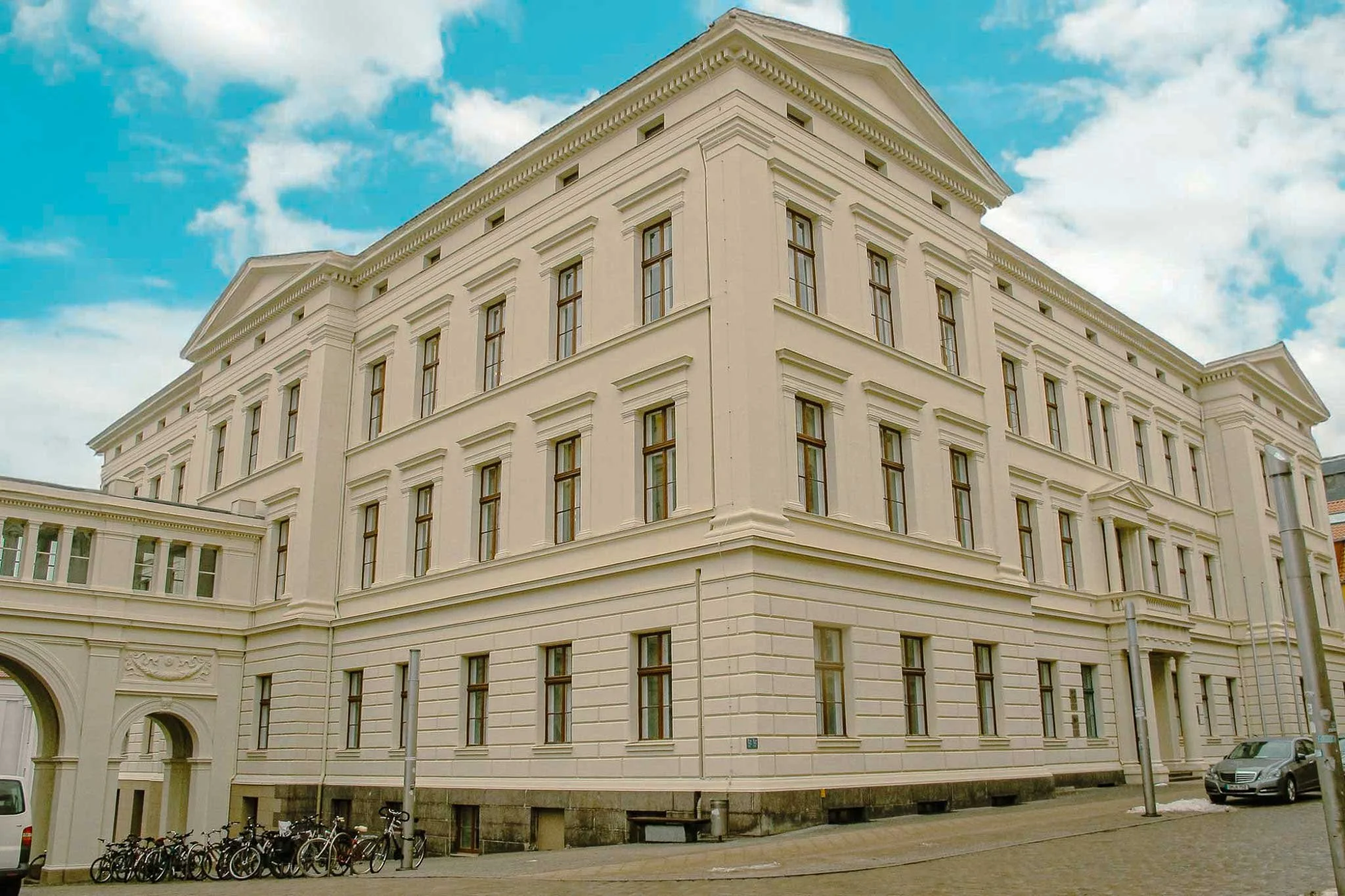 Ein weißes, mehrstöckiges Gebäude mit klassischen architektonischen Elementen, mehreren Fenstern und einem blauen Himmel mit einigen Wolken im Hintergrund.