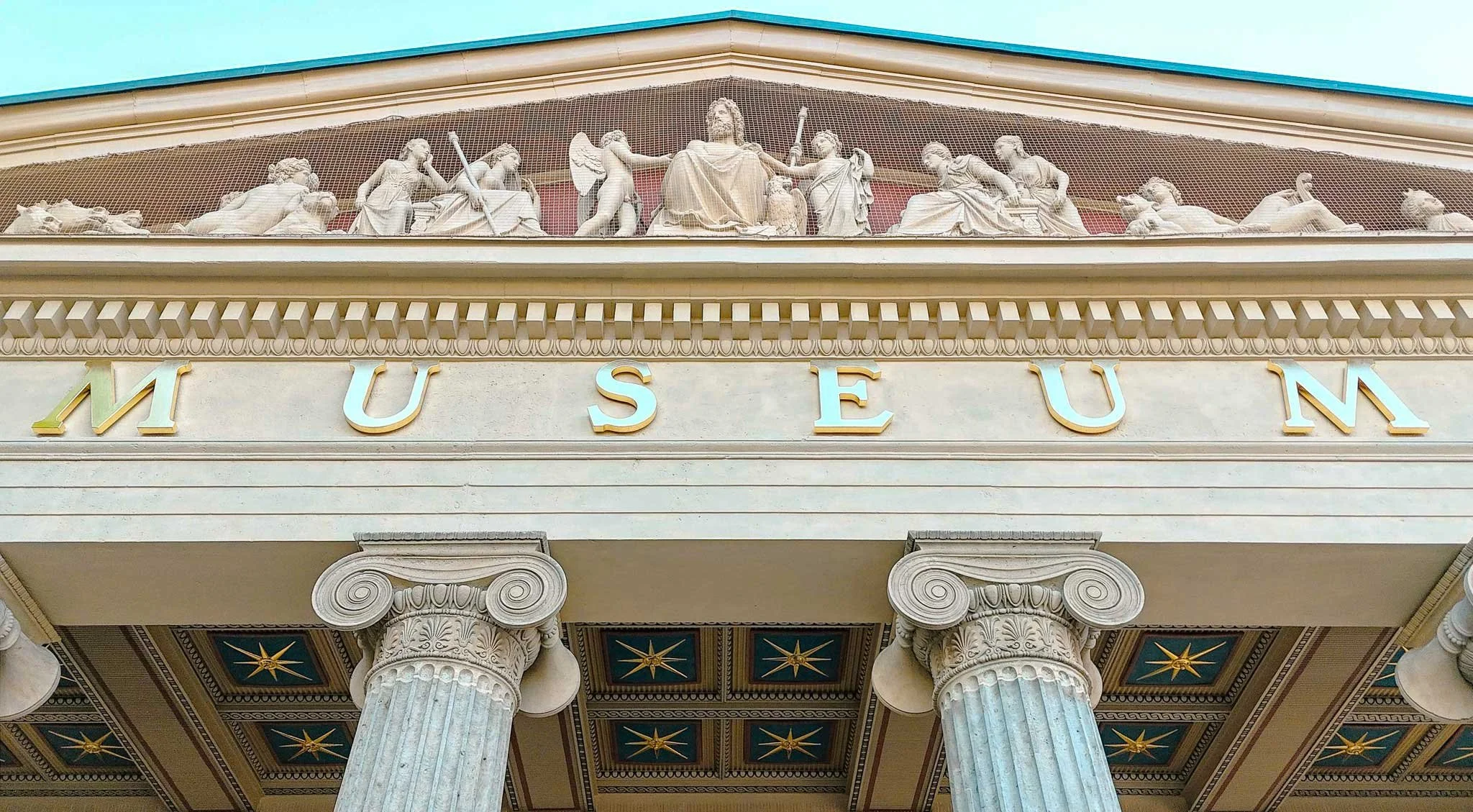 Ansicht der Fassade eines Museums mit klassischen Säulen und einer Friese mit Skulpturen und Figuren. Das Wort 'MUSEUM' ist in großen Buchstaben oberhalb der Säulen sichtbar.