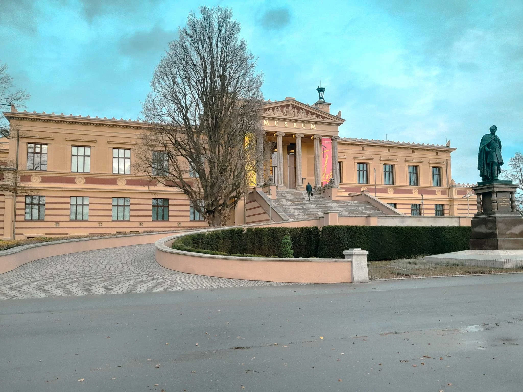 Das Bild zeigt ein neoklassizistisches Museum mit Säulenfront und einem Trauerschmuck, umgeben von Bäumen und einer Statue im Vordergrund.
