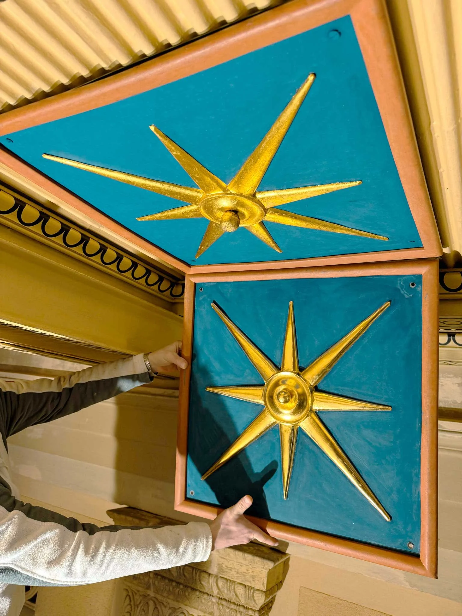 Zwei Hände halten eine bemalte Holzplatte mit einem goldenen Sternschnuppen-Design vor einem Raum mit gelben und beige Wänden.