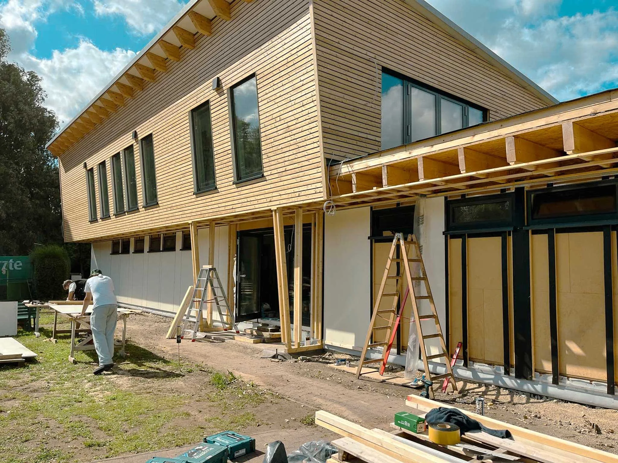 Baustelle eines modernen Hauses mit Holzfassade, Bauarbeiter arbeiten an Fenstern und Terrassenbereich, Gerüste und Werkzeuge sind sichtbar.