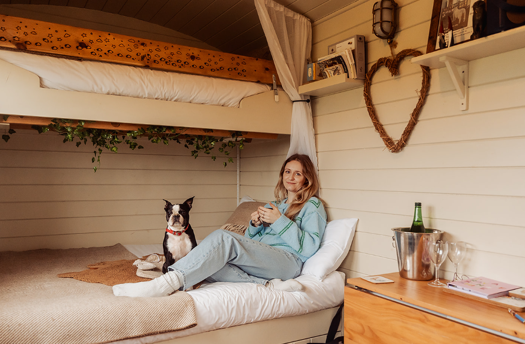 A woman sitting on a bed in a cozy wooden room with a dog next to her. She is holding a mug and smiling. The bed has beige and white bedding, and there is a loft bed above with a brown blanket. The room has shelves with books and decor, including a heart-shaped wreath on the wall and a table with a bottle of wine, glasses, and notebooks.
