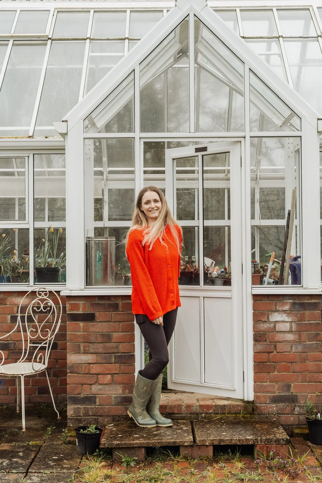 A woman in a red sweater, gray pants, and green rubber boots standing in front of a greenhouse with a brick wall. The woman is smiling, and the greenhouse has large glass panels and a door. There is a white metal garden chair and some small potted plants on the ground.
