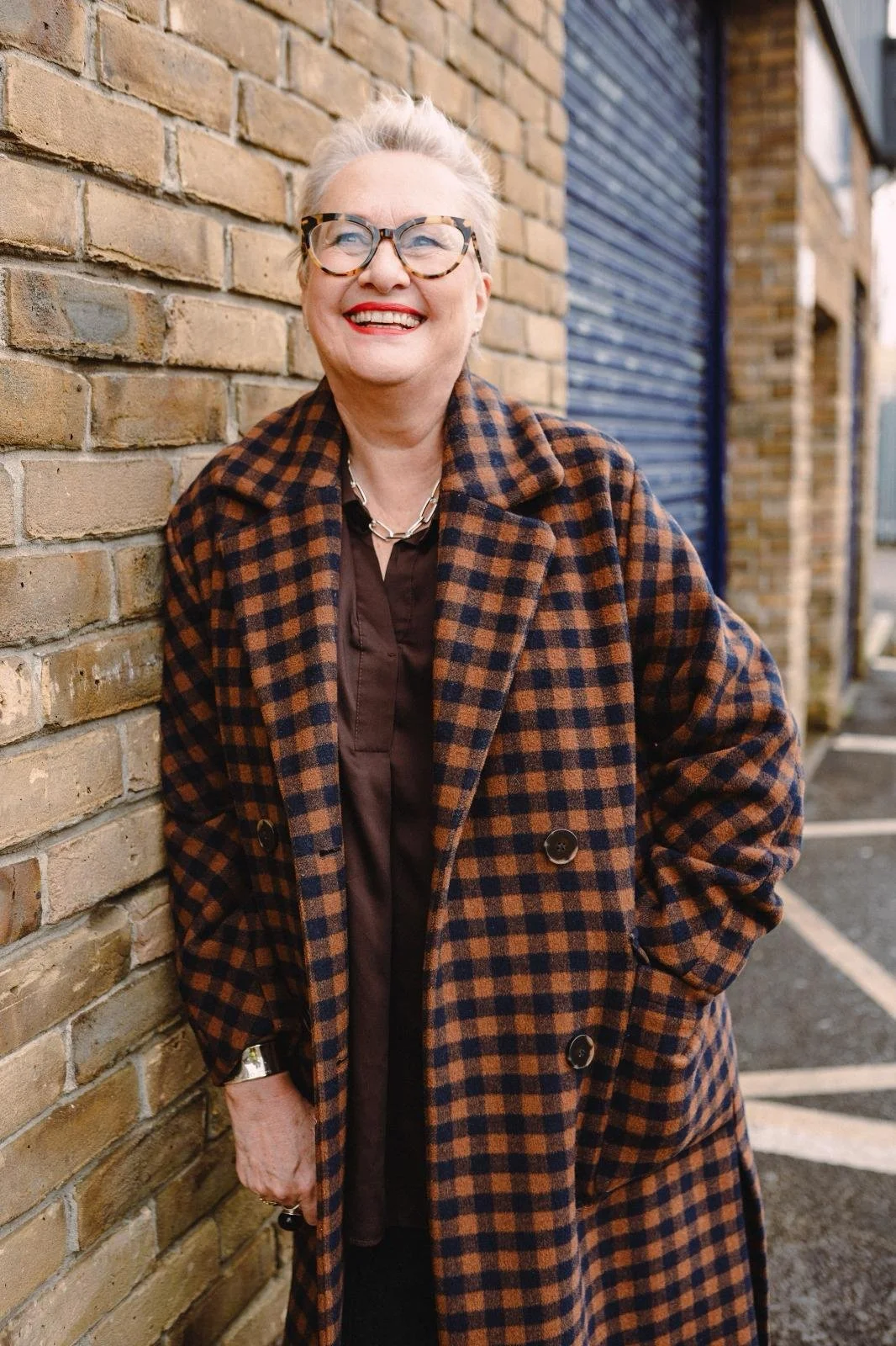 A cheerful woman with short gray hair, glasses, red lipstick, wearing a brown and orange checked coat and standing against a brick wall on an urban street.