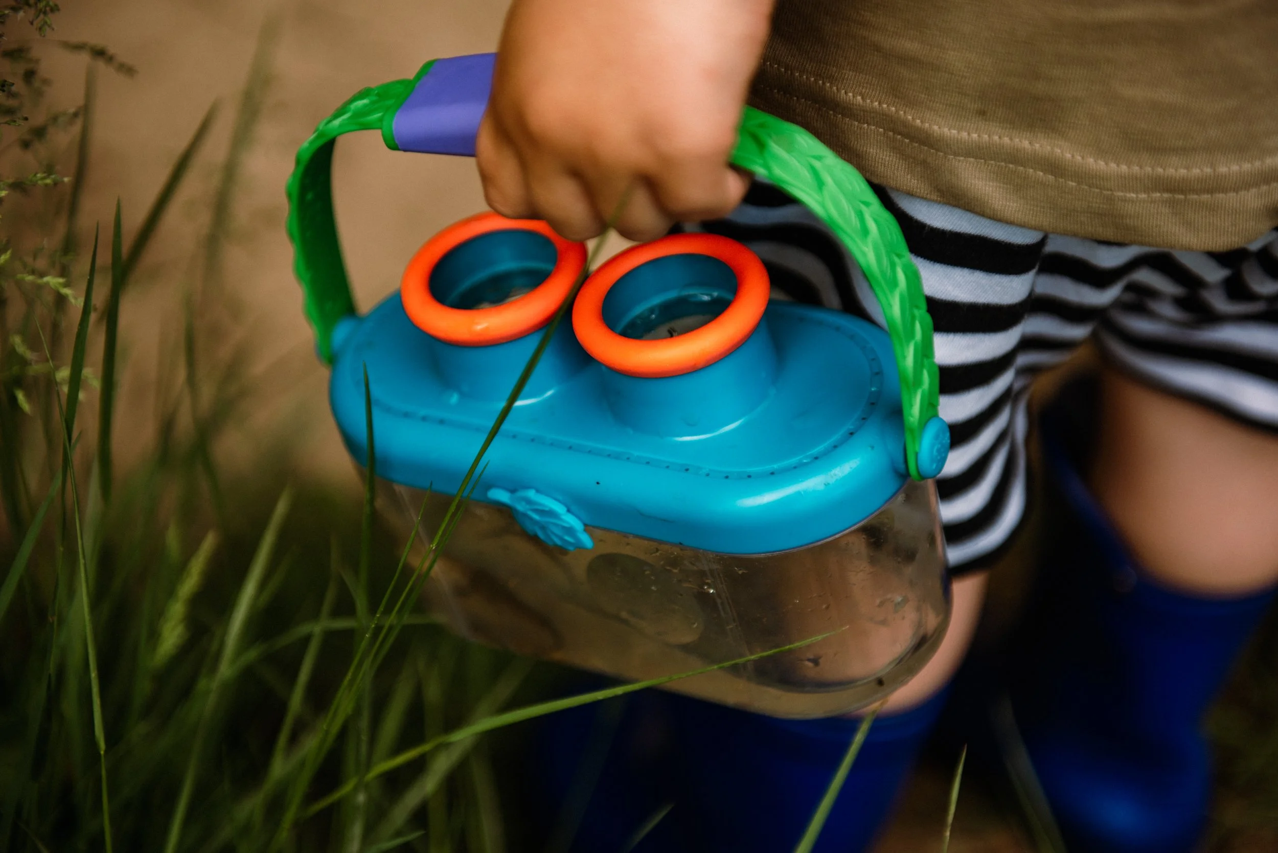 A child holding a colorful toy with a green handle and orange rings, with grass and a blurred background.
