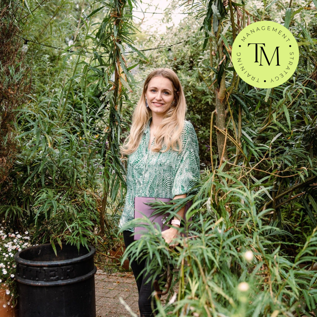 A woman with long blonde hair standing in a lush garden with green plants. She is smiling and holding a purple folder, wearing a green patterned blouse. There is a black flower pot near her and a circular logo with the words 'Management,' 'Strategy,' 'Training,' and 'Turning' in the upper right corner.