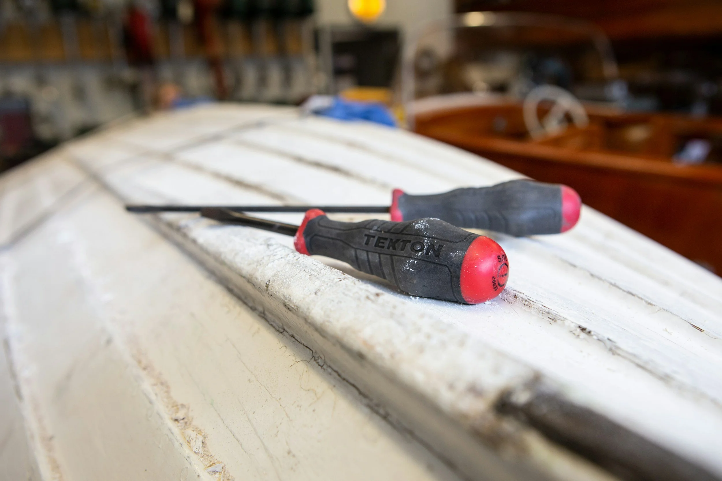 Close-up of two screwdrivers with red and black handles resting on a white painted surface, possibly a windowsill or countertop, with a blurred background of workshop or garage tools and furniture.