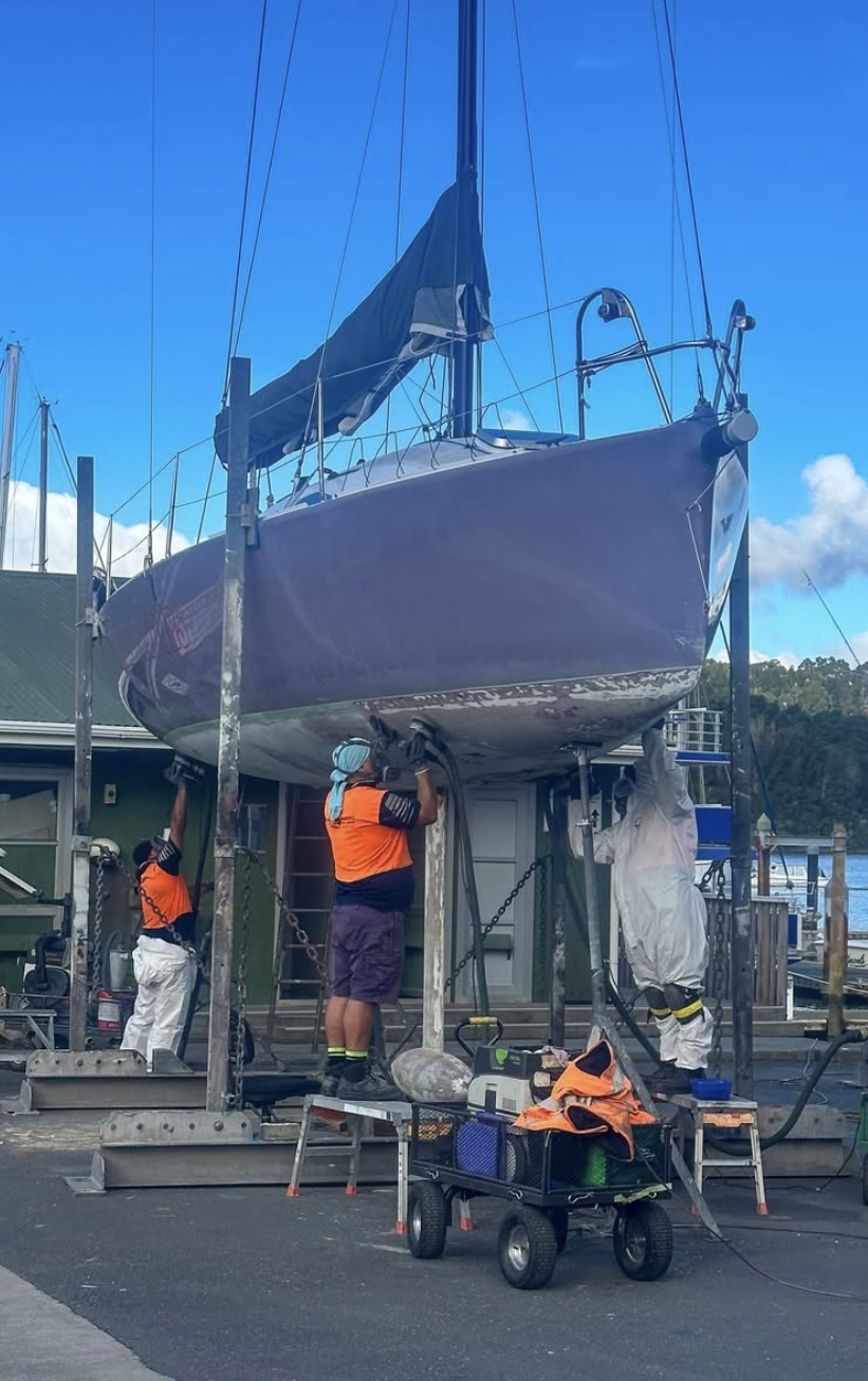 Three workers are repairing or cleaning the bottom of a sailboat that is lifted up on a boat stand at a marina. They are using tools and wearing safety gear, with a clear blue sky in the background.