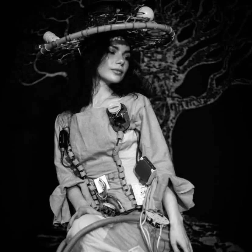 Donna in abito lungo con accessori tecnologici, indossa un grande cappello decorato con fili e circuiti elettronici, in uno sfondo oscuro con un albero distorto.