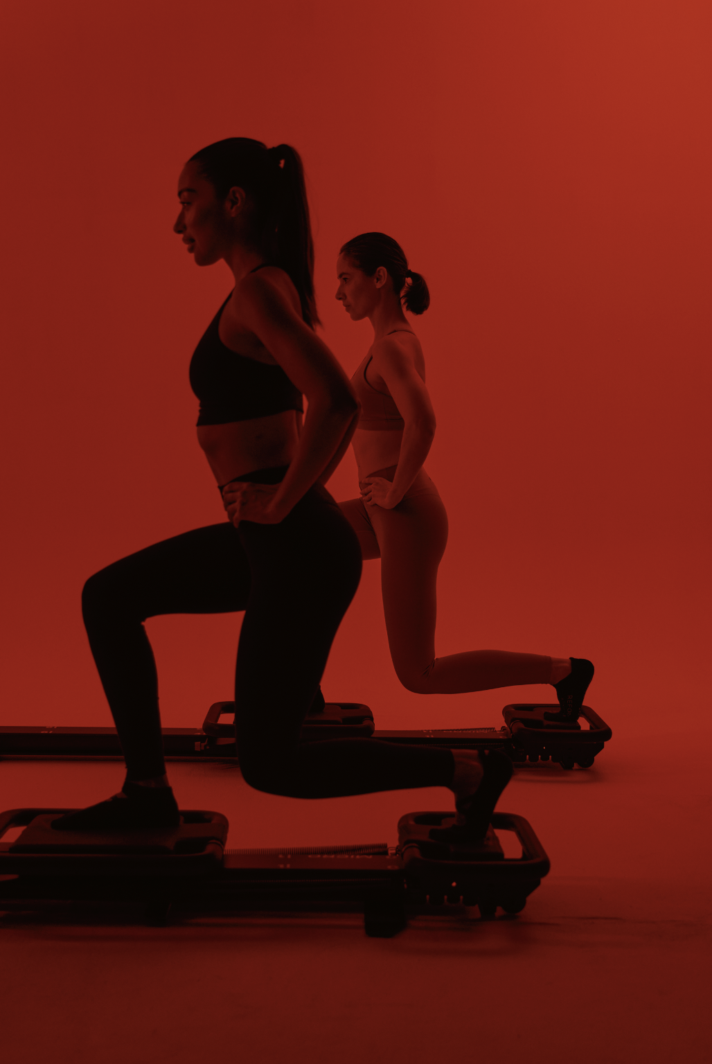 Two women performing exercises on step platforms in a fitness studio with a red background.