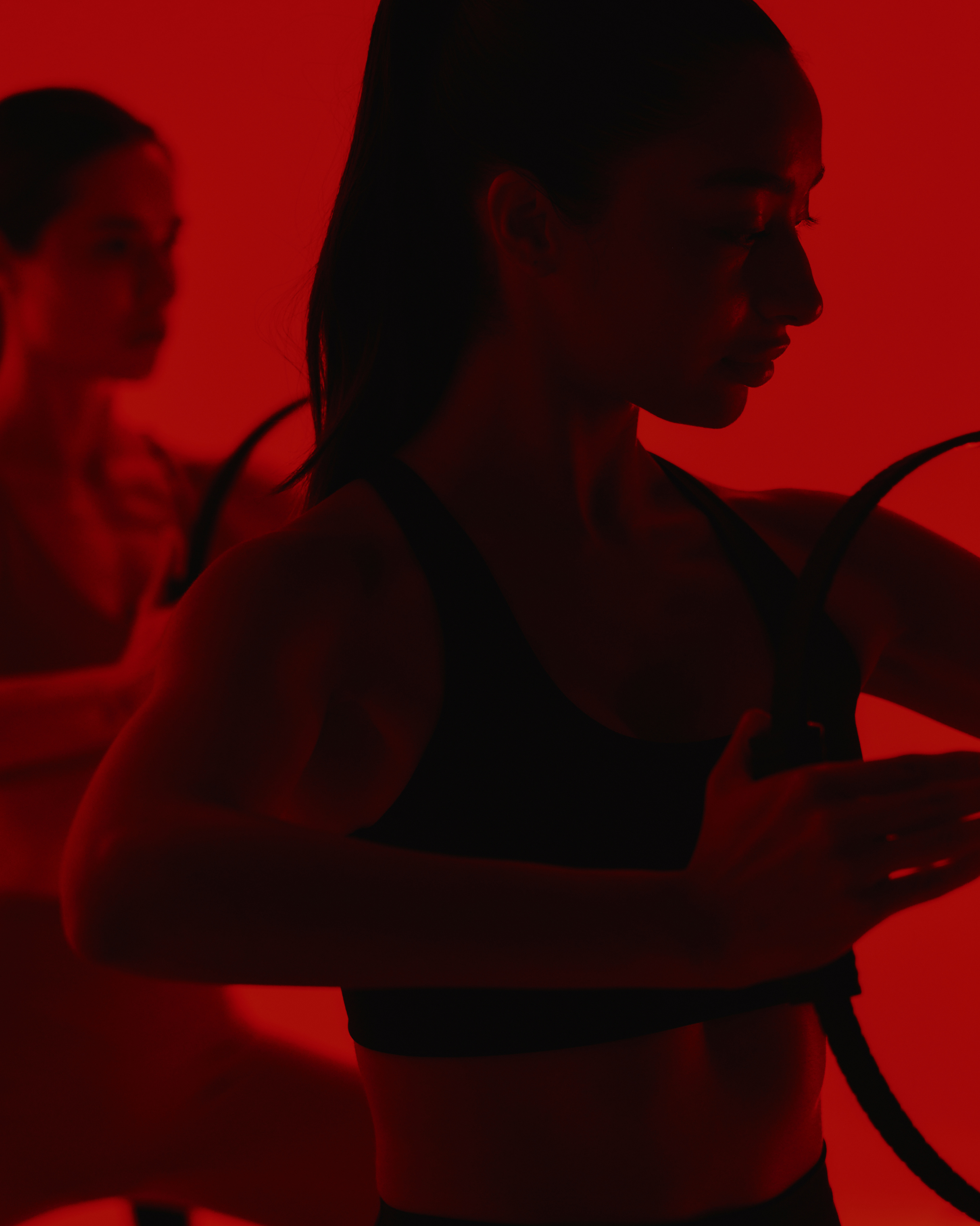 Silhouettes of two women in dark athletic wear against a red background. One woman is in the foreground, looking slightly downward, and the other woman is in the background, slightly out of focus.
