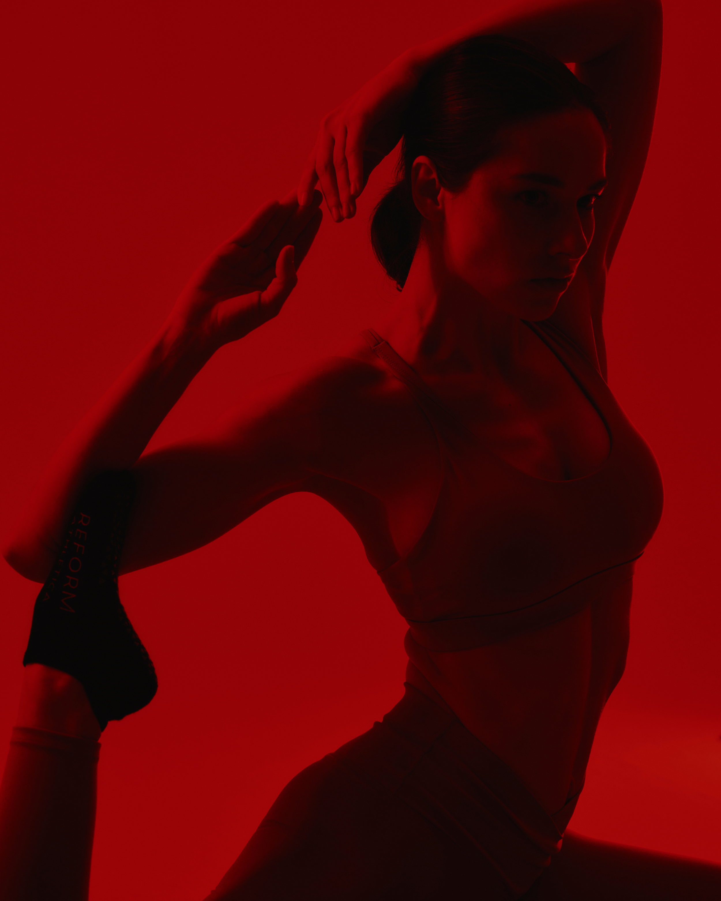 A woman stretching with one arm bent behind her head and the other hand touching her forehead, silhouetted against a red background.