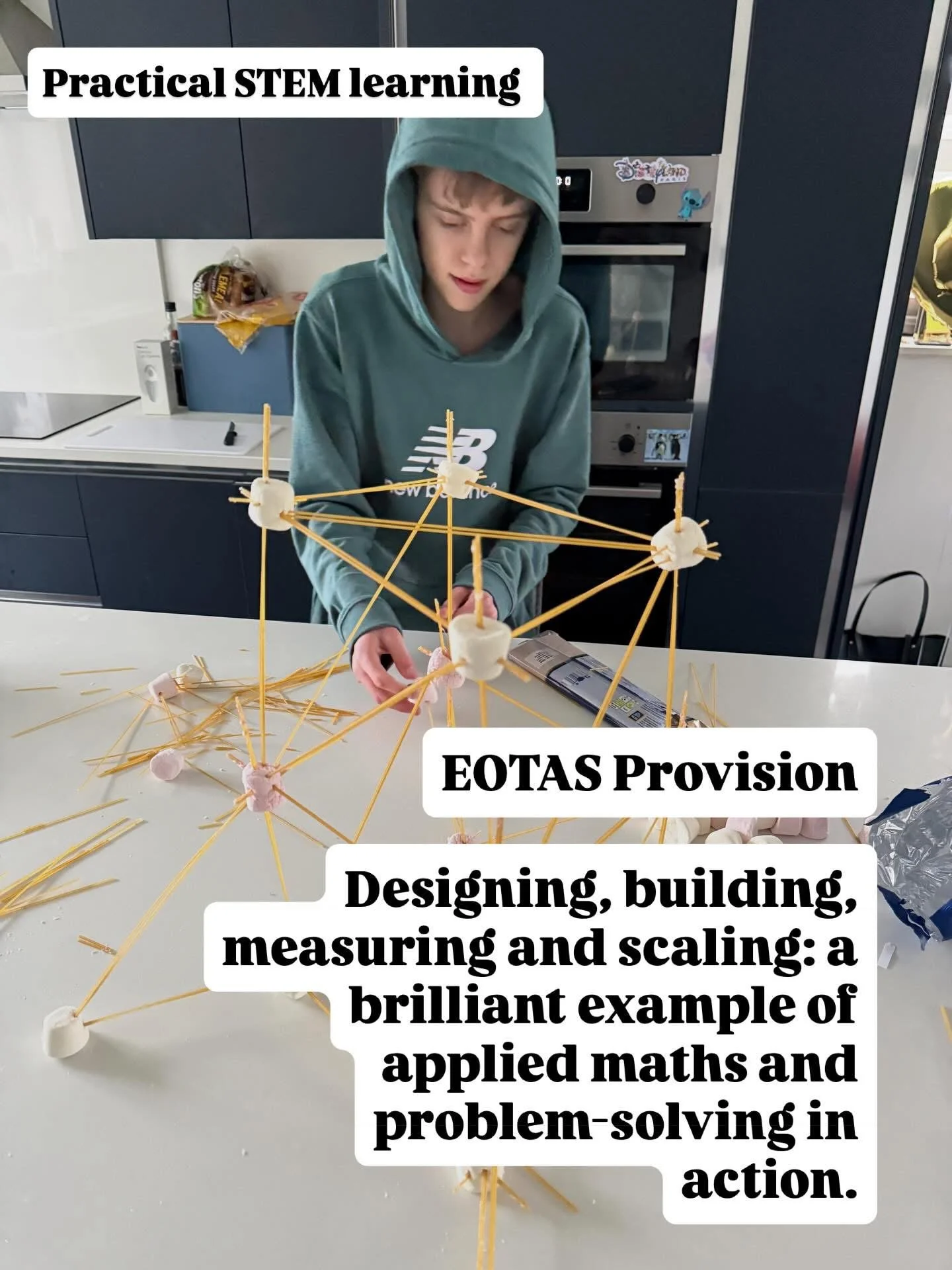 A young person in a green hoodie is working on a STEM project involving a structure made of spaghetti and marshmallows on a white table. The background shows a kitchen with cabinets and appliances.