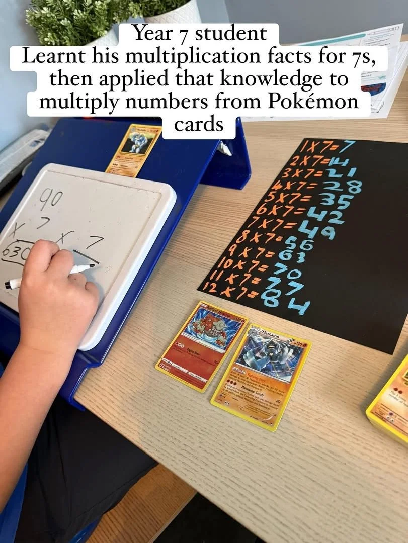 A young student practicing multiplication facts with Pokémon cards and handwritten equations on a whiteboard, displayed on a desk with a black paper showing multiplication tables.