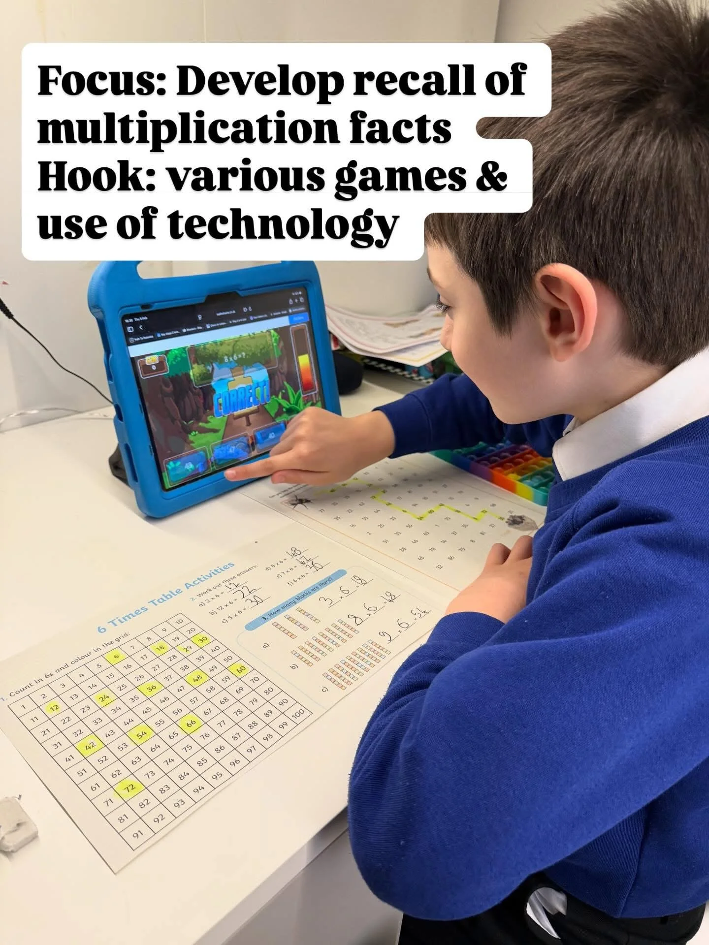 A young boy in a blue school uniform is sitting at a desk, using a tablet to play an educational game about multiplication. On the desk, there is a colorful multiplication chart and worksheets with math exercises.