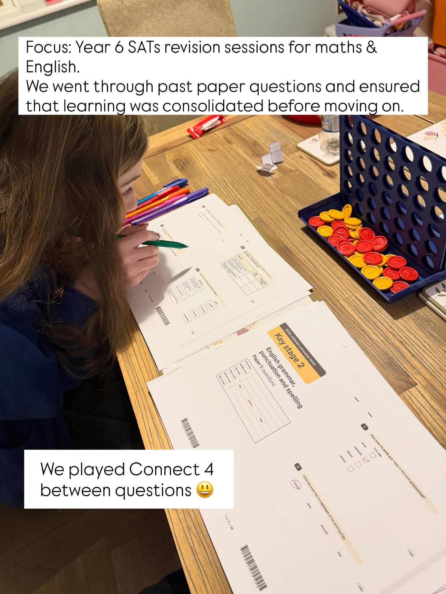 A girl in a dark blue shirt is sitting at a wooden table, working on a worksheet related to English grammar, punctuation, and spelling. There are various colored pens, a water bottle, and a game with red and yellow tokens on the table. Some questions