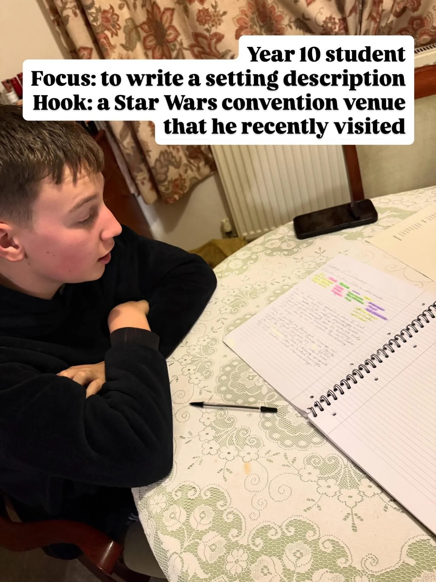 A young boy sitting at a table with a notebook, some highlighted notes, a pen, and a smartphone. He appears to be studying or doing homework in a room with floral curtains and a radiator.
