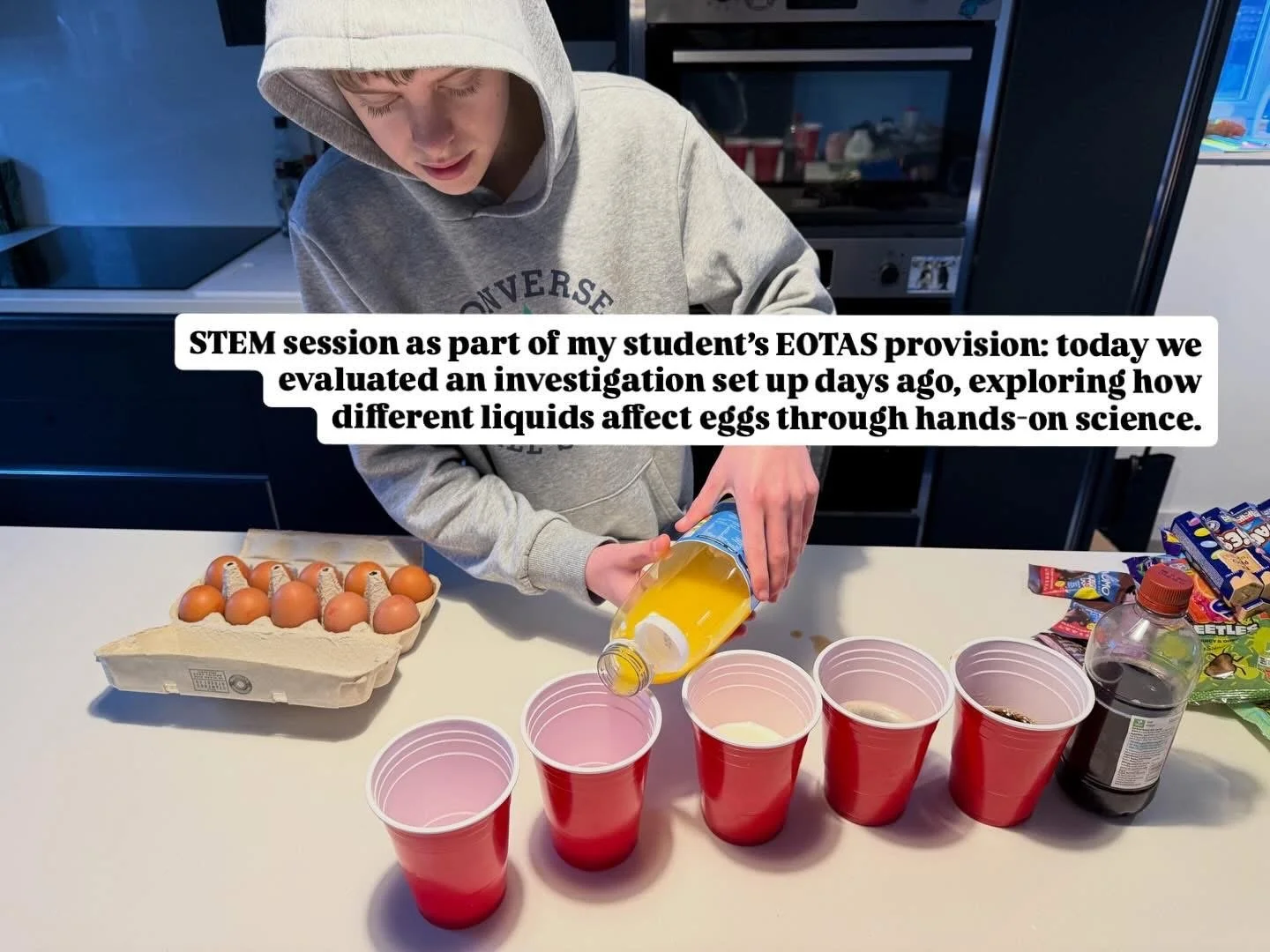 A student pouring yellow liquid from a bottle into pink cups on a white table, with a carton of eggs and various candy packets nearby, as part of a science experiment.
