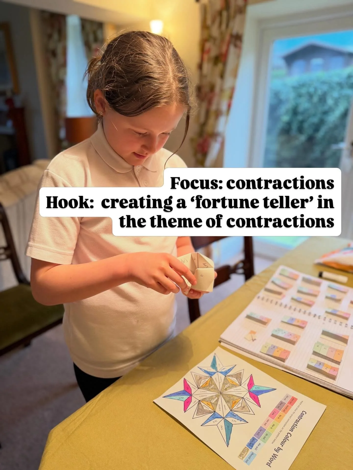 A young girl with brown hair and a beige polo shirt is examining a paper fortune teller and color-coded charts laid out on a yellow table in a well-lit dining room with floral curtains and a large window showing an outdoor scene.