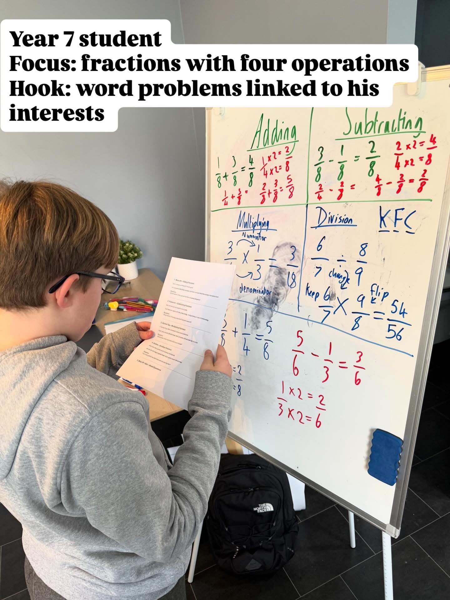 A boy with glasses and a gray hoodie studying something on a piece of paper. He is standing next to a whiteboard with fractions written on it, and a backpack is on the floor nearby.