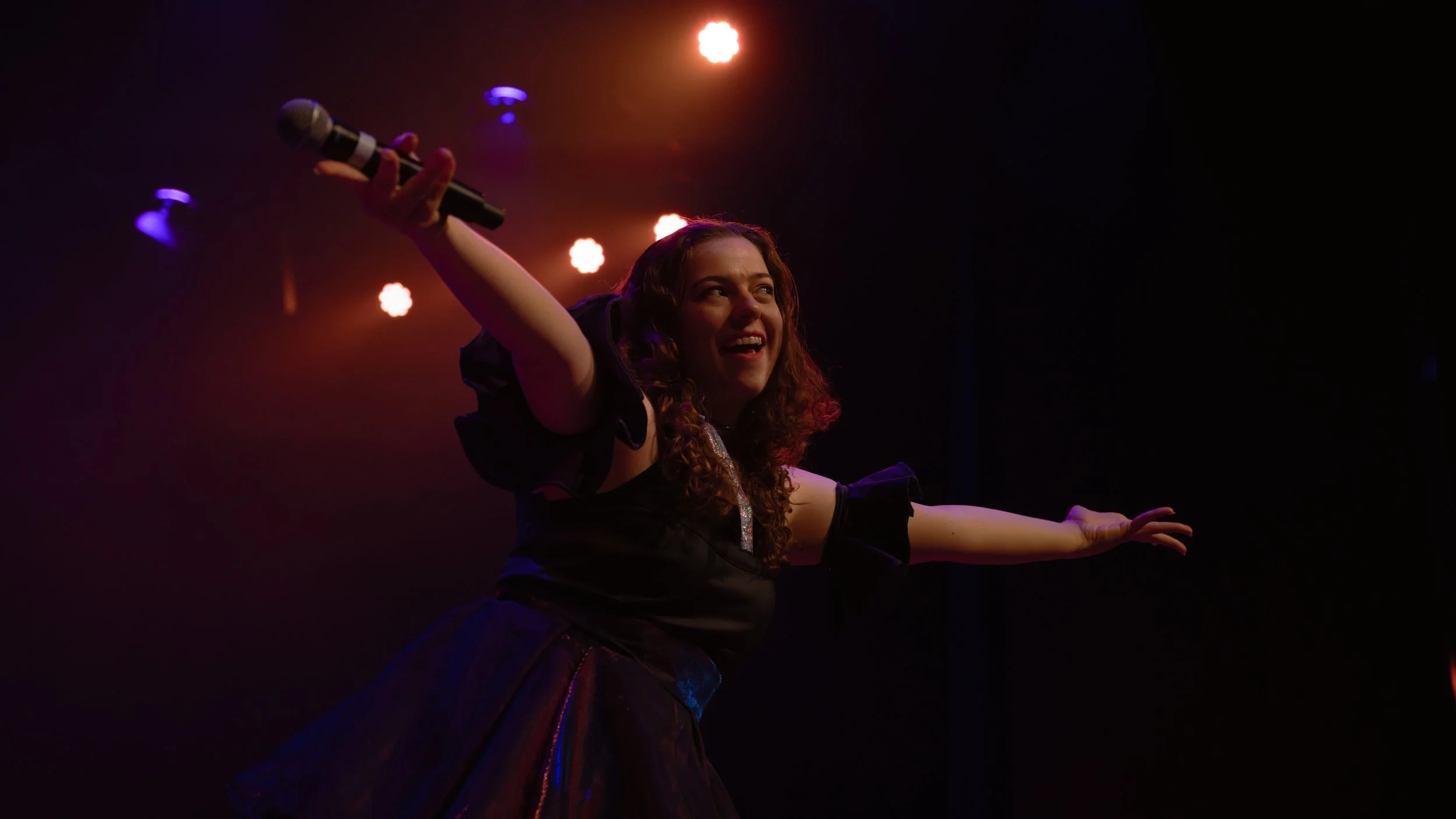 A woman performing on stage, holding a microphone in her right hand, smiling and extending her left arm outward against a dark background with stage lighting.