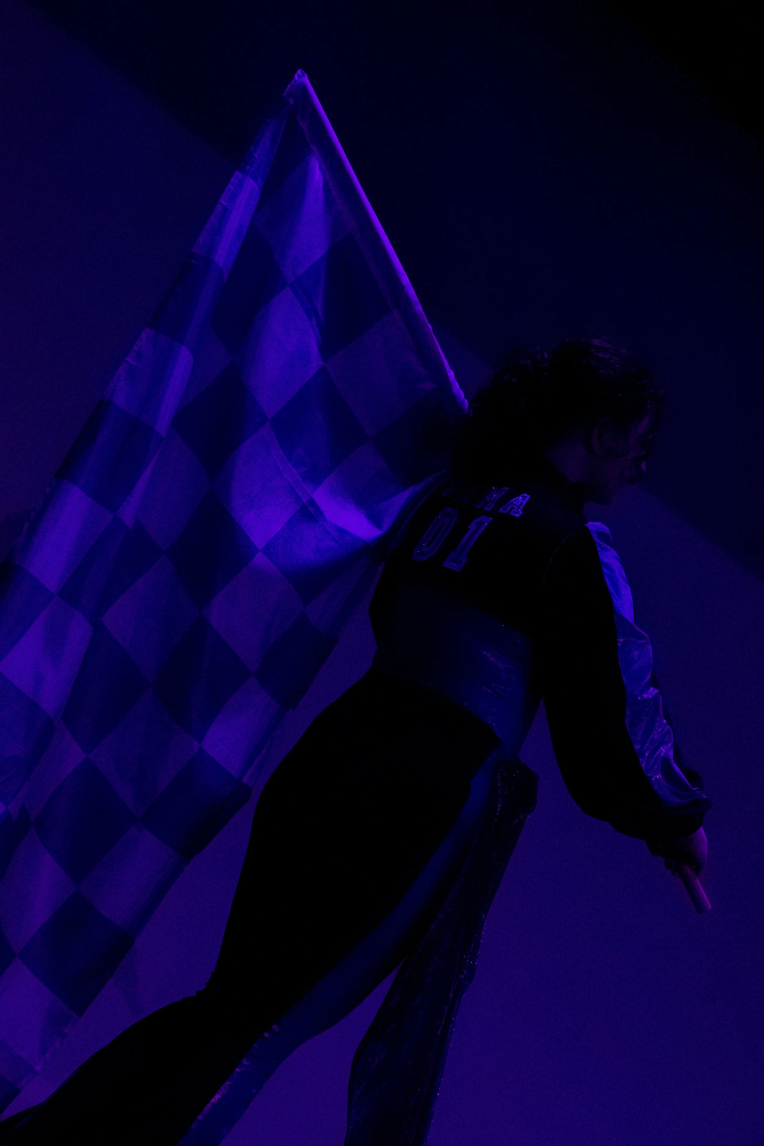 Person with curly hair and sunglasses leaning against a checkered flag, illuminated by purple and blue lighting in a dark environment.