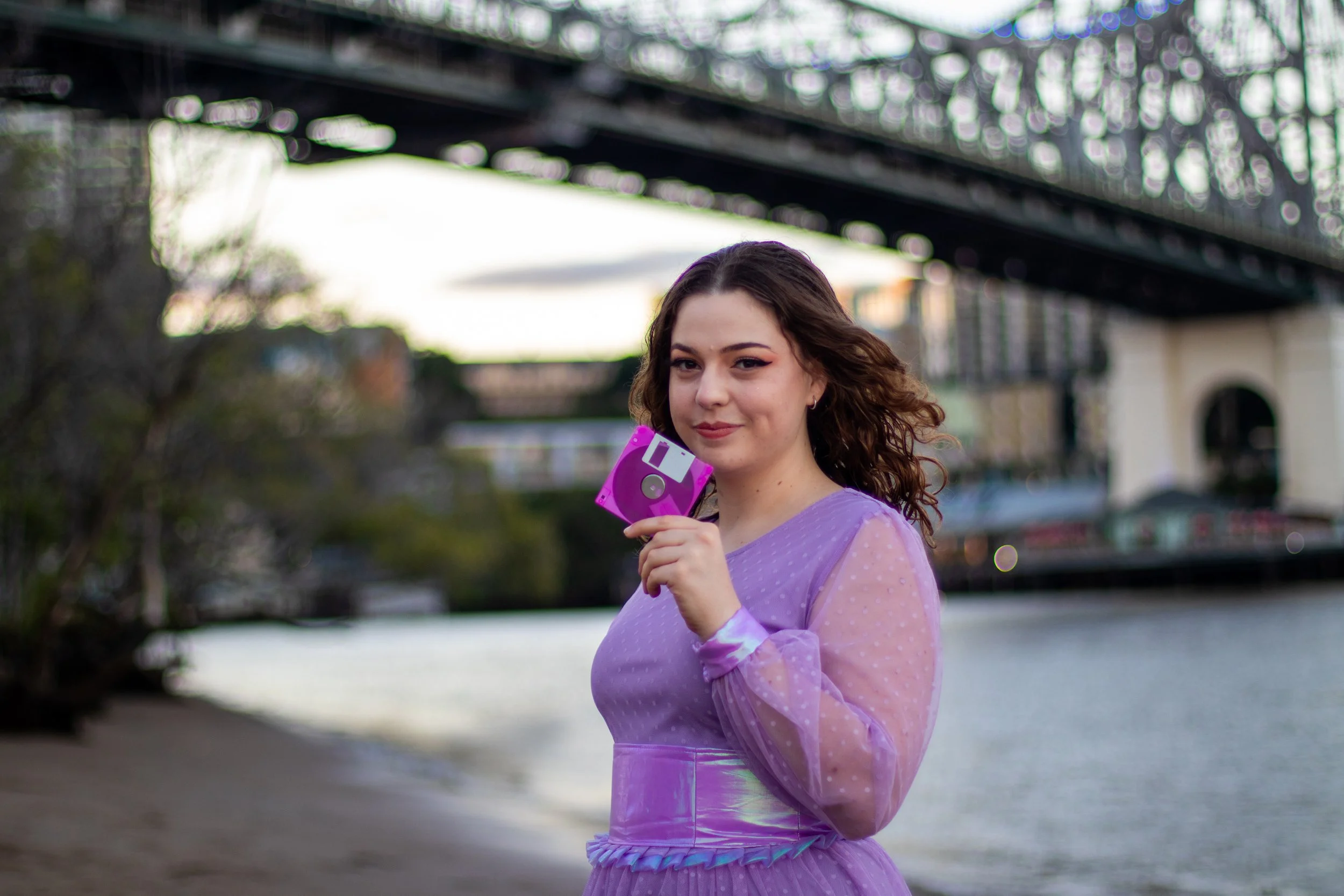 A young woman with wavy brown hair wearing a purple dress with sheer polka-dot long sleeves, holding a pink and gray floppy disk near her face outdoors near a river with trees and a bridge in the background.