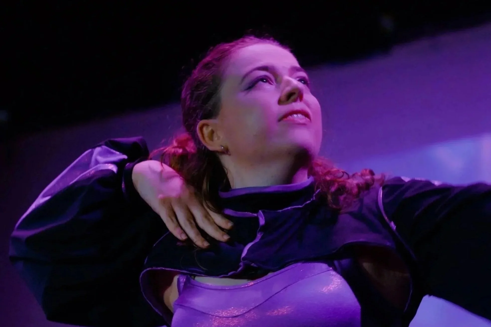 A woman with reddish-brown hair is in a dance pose in a dimly lit environment with purple and blue lighting, wearing a dark jacket and white pants.