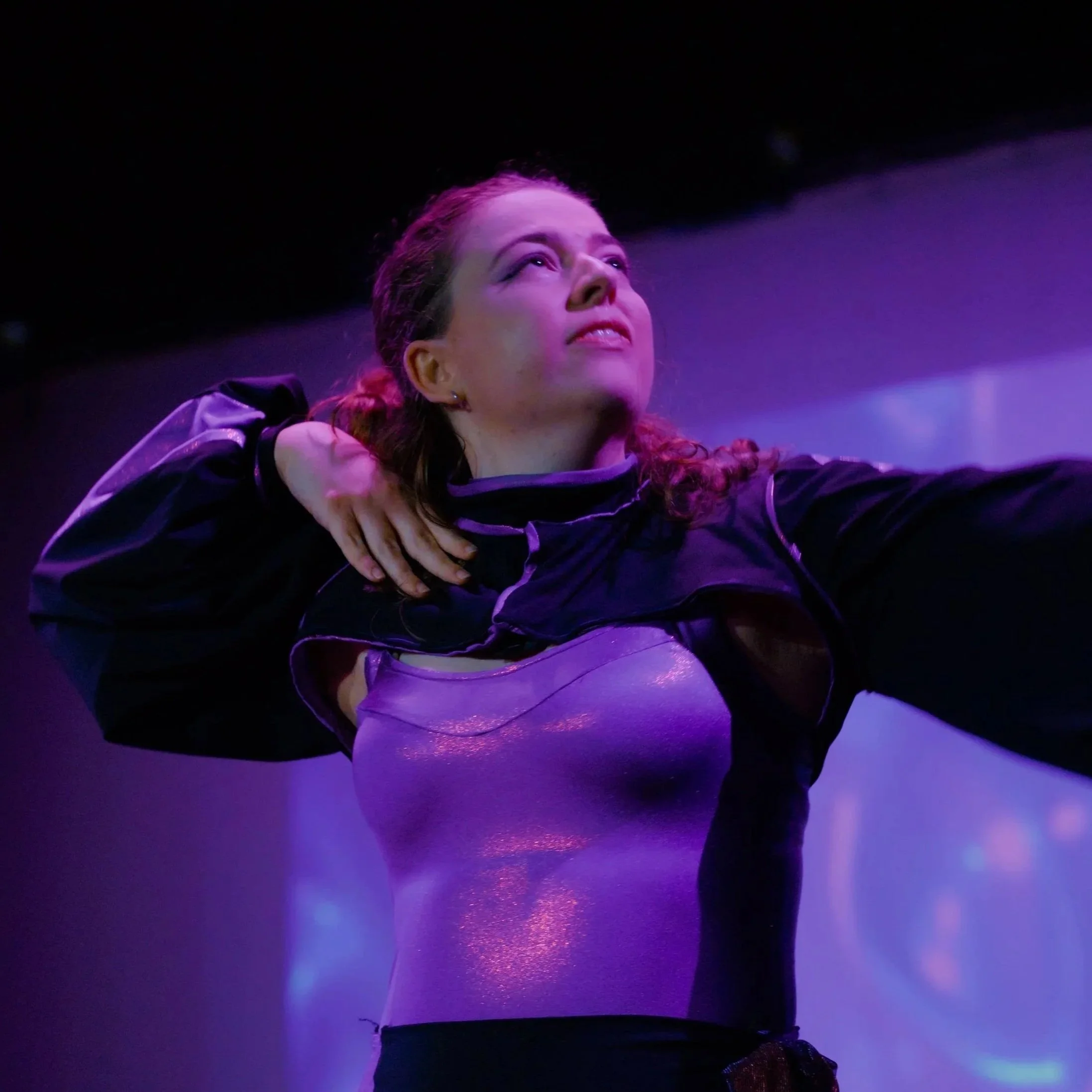 A woman with reddish-brown hair is in a dance pose in a dimly lit environment with purple and blue lighting, wearing a dark jacket and white pants.
