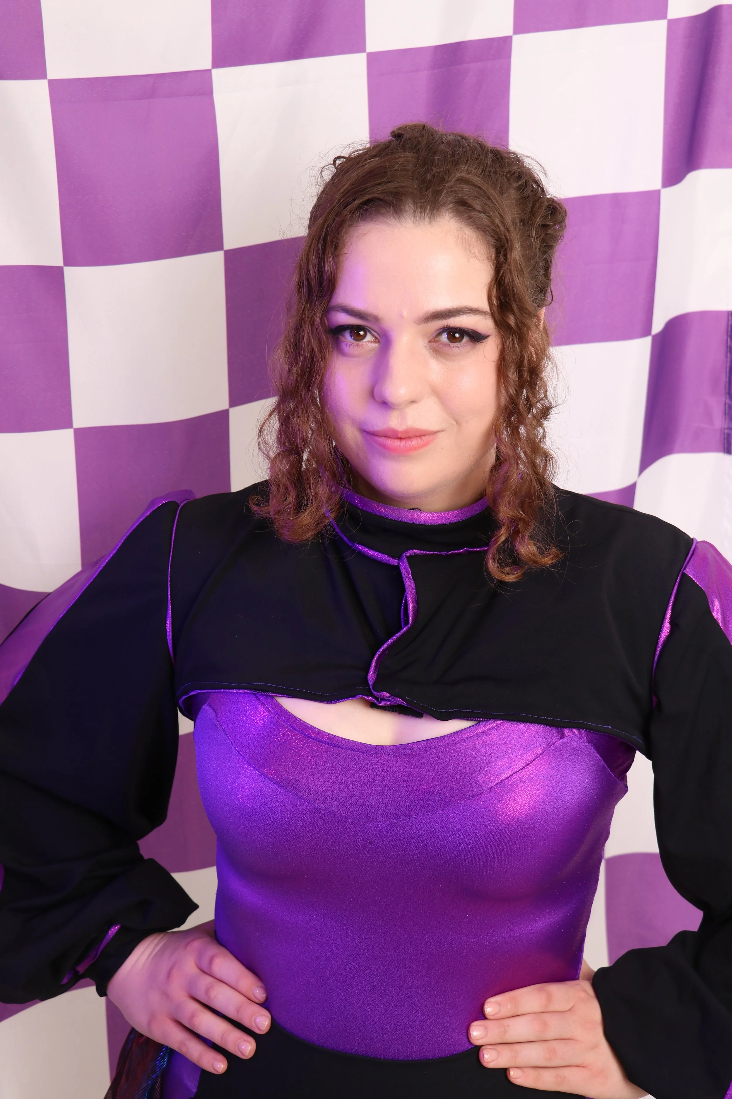 A woman with curly brown hair wearing a purple and black outfit, standing in front of a white and purple checkered background.