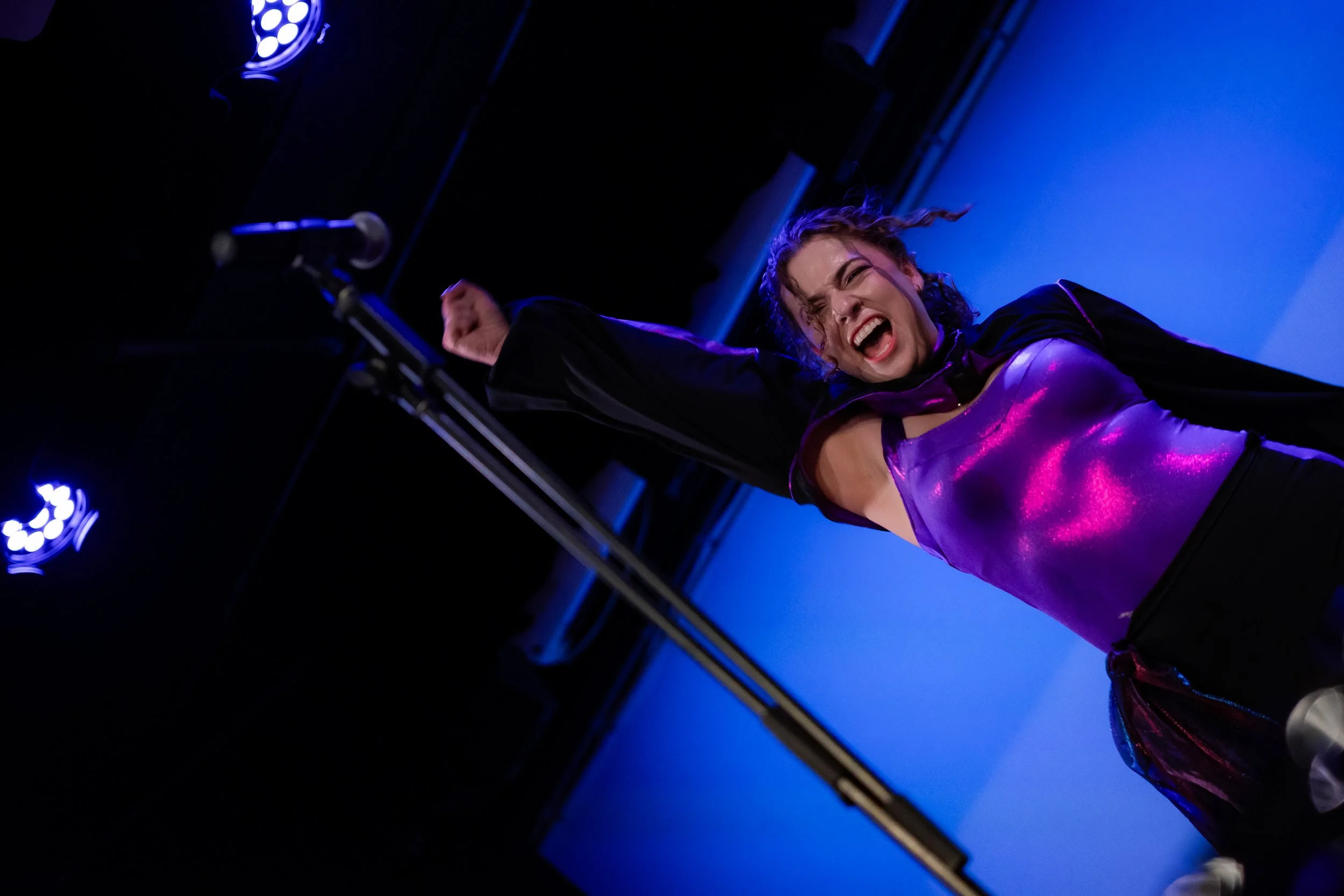 Young woman passionately singing or performing on stage, illuminated with purple and blue stage lights.