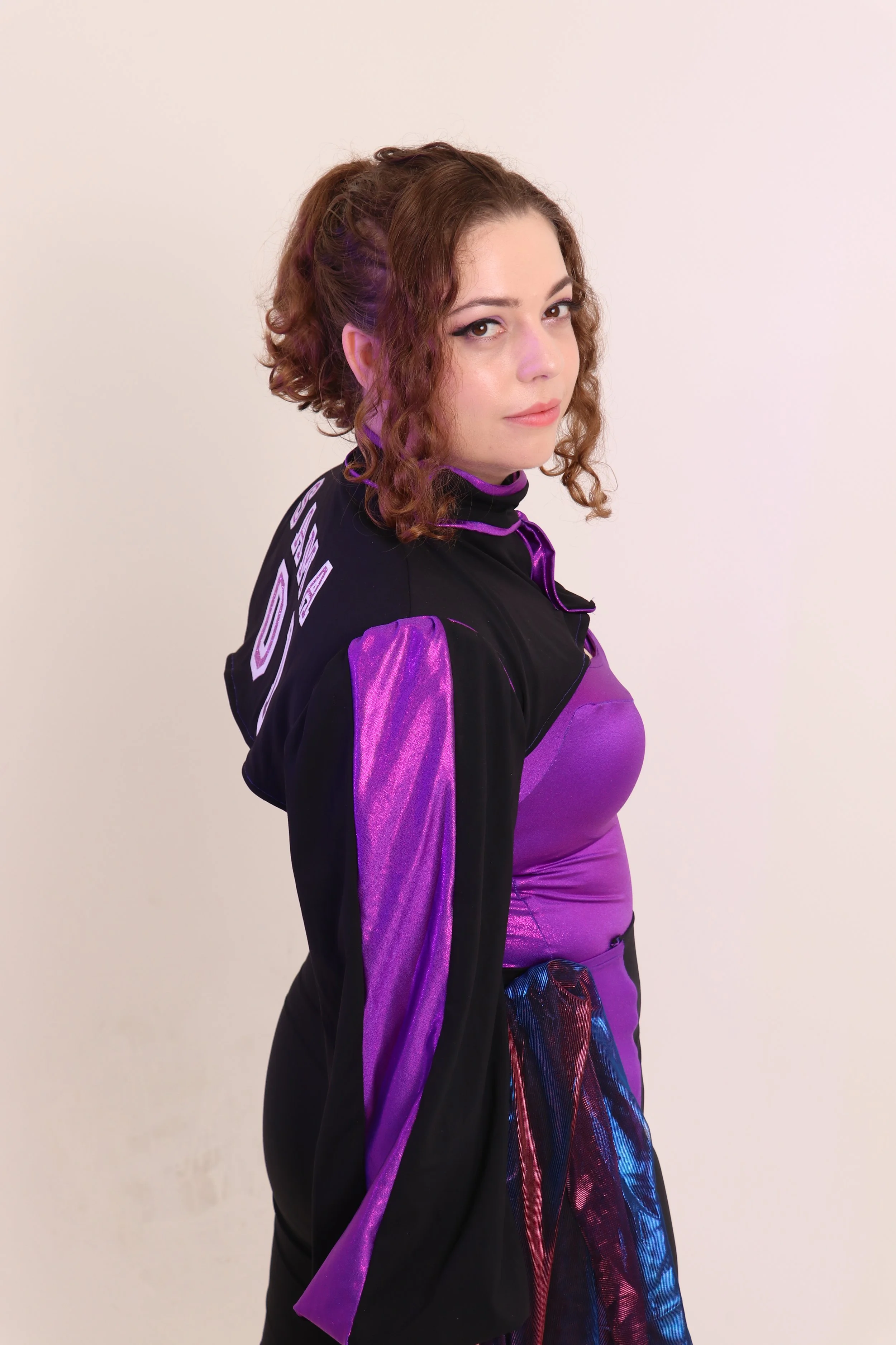 Woman with short curly brown hair wearing a black and purple jacket and a purple dress, standing against a plain white background.