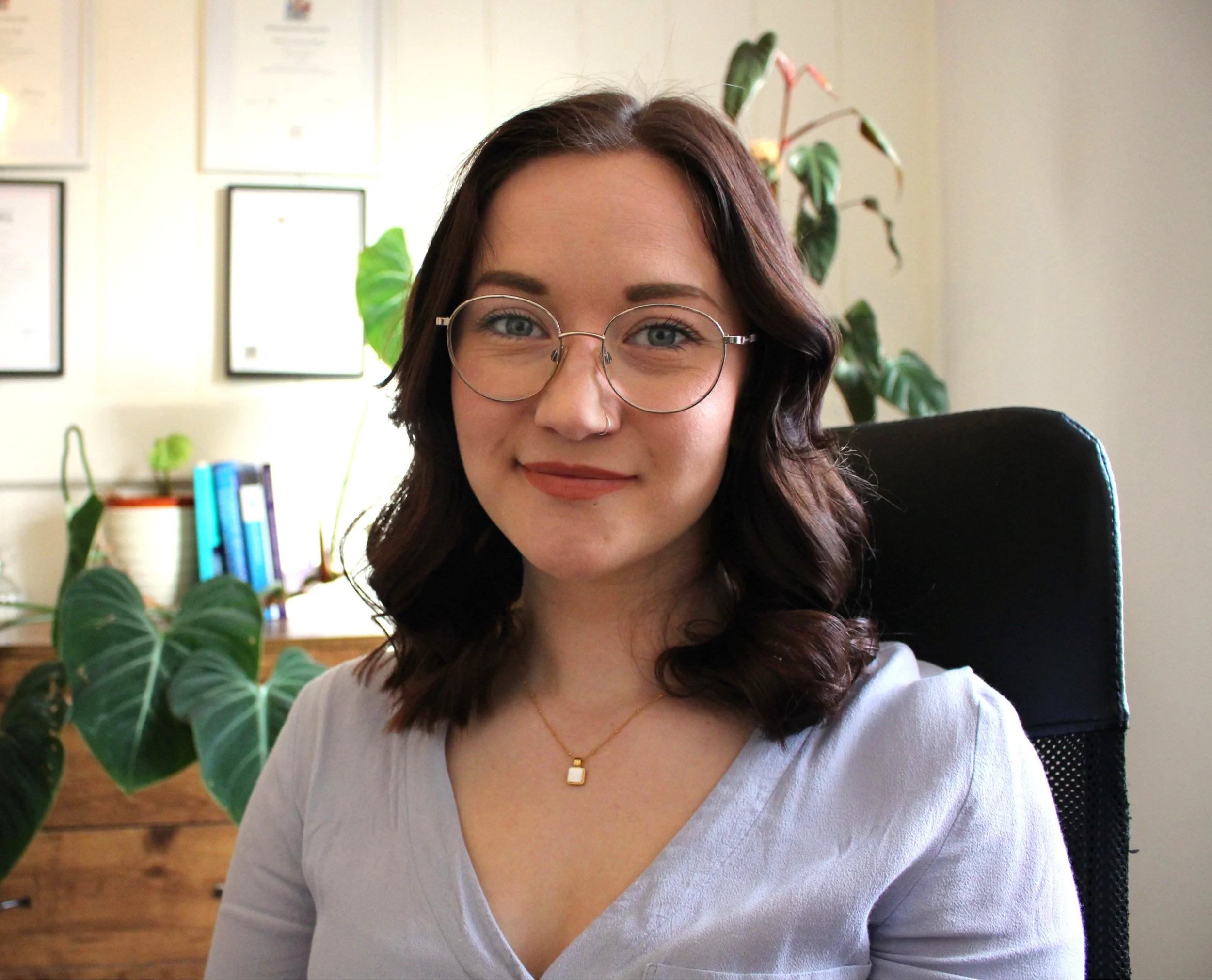 A woman with dark wavy hair and glasses sitting in an office, with certificates on the wall and plants in the background.