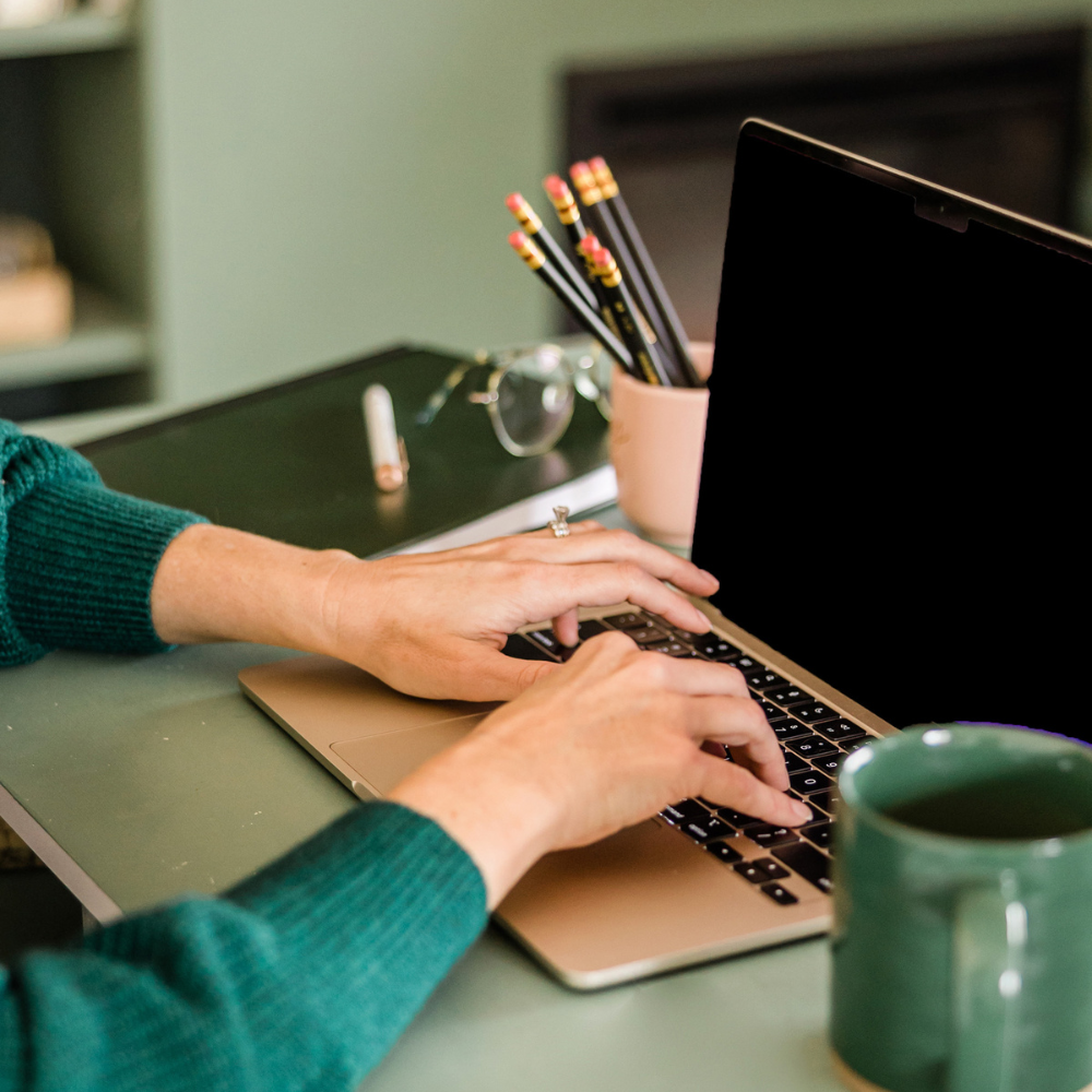Person typing on a laptop keyboard at a desk with a cup, glasses, pencils, and a notebook.