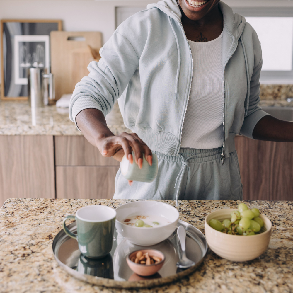 A person in a white hoodie and sweatpants pouring milk into cereal in a white bowl on a granite kitchen countertop, with a bowl of green grapes, a mug, and a small bowl of nuts nearby.