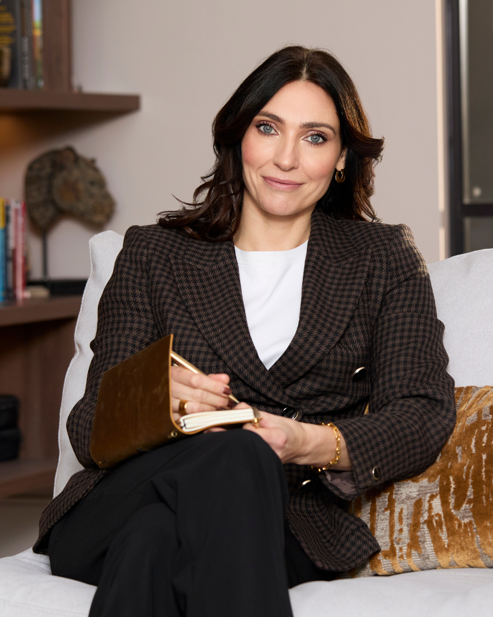 A woman with dark hair and blue eyes, wearing a brown checkered blazer and white blouse, sitting on a white sofa with a book and smiling at the camera in a cozy, modern living room.
