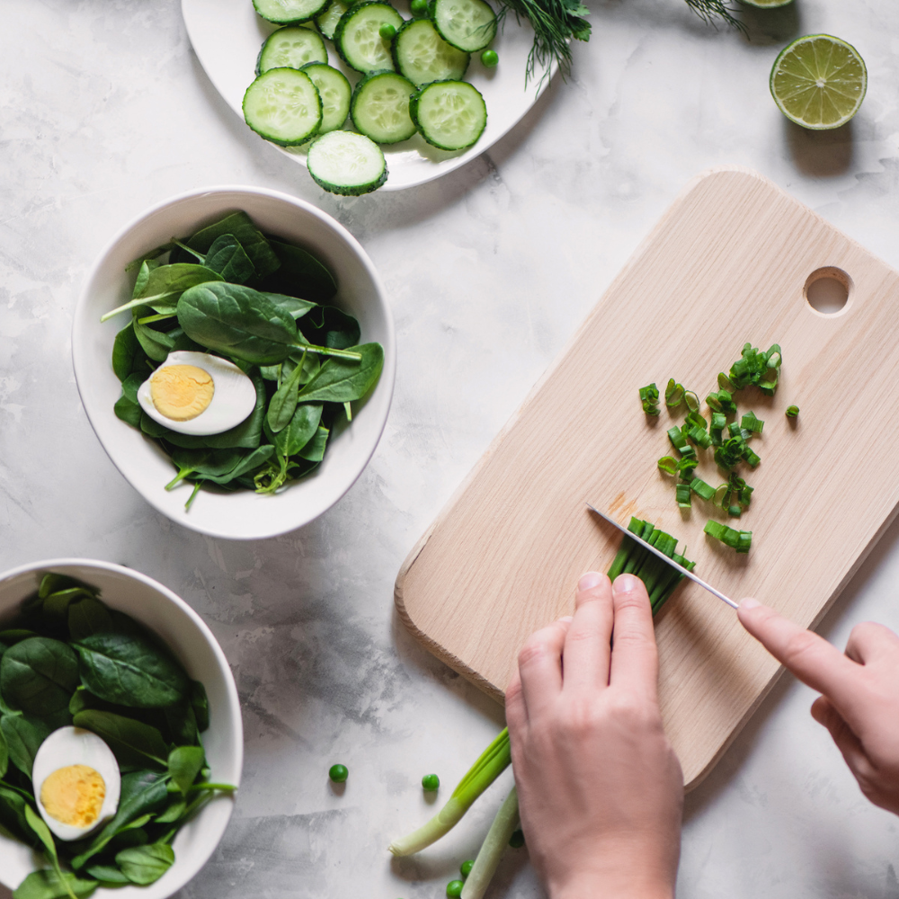Person chopping green onions on a wooden cutting board surrounded by bowls of spinach salads with boiled eggs, sliced cucumbers, green peas, and a halved lime.