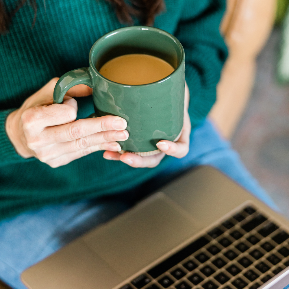 Person holding a green mug of coffee or tea while sitting near a laptop.