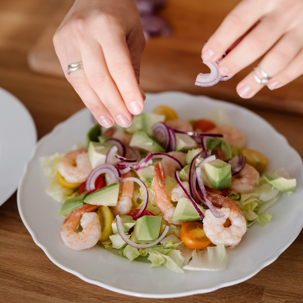 Hands adding sliced red onion to a shrimp and avocado salad with cherry tomatoes on a white plate.