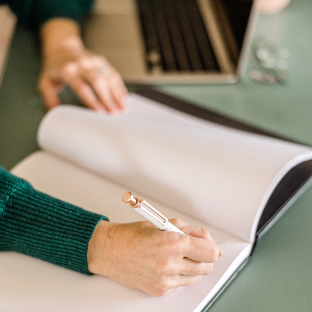 A person with a green sweater writing in a blank notebook with a white pen with a pink top, on a green desk with a container of small coins and a book in the background.