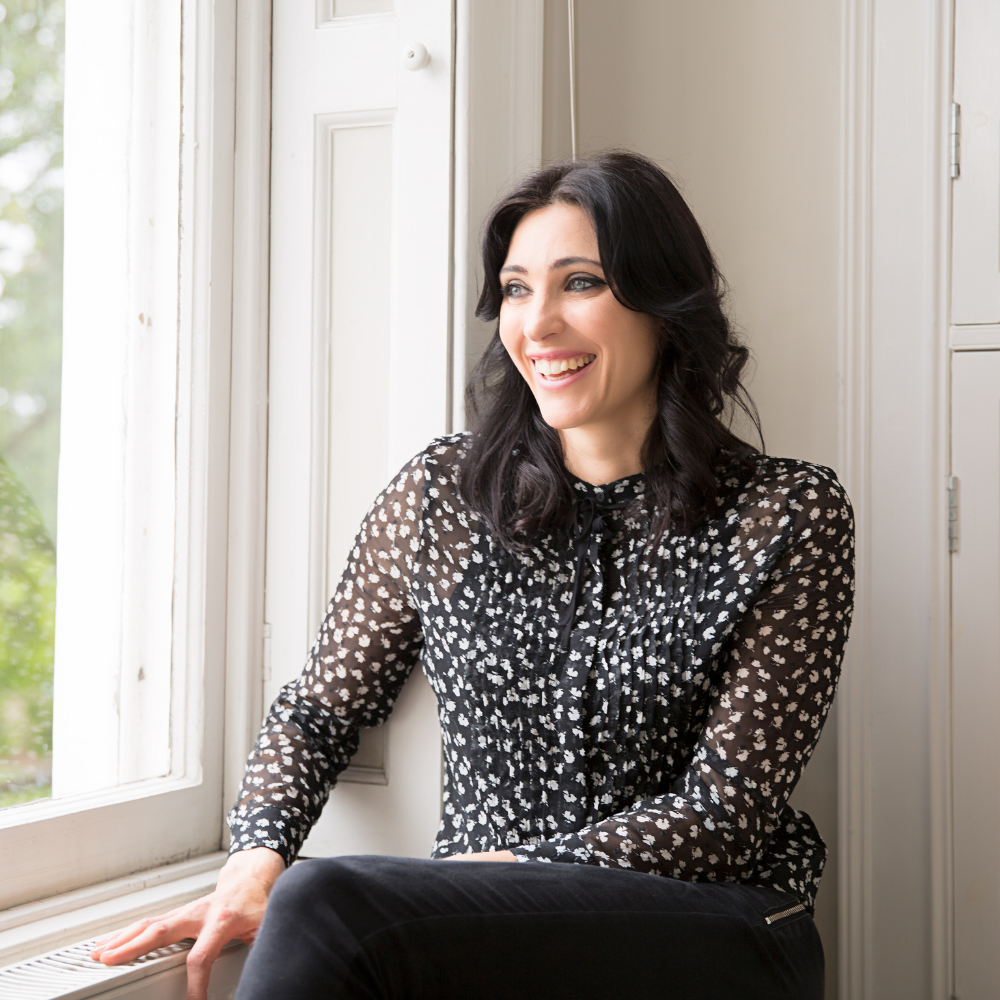 A woman with black wavy hair and light skin sitting by a sunlit window, smiling and wearing a black and white patterned blouse.