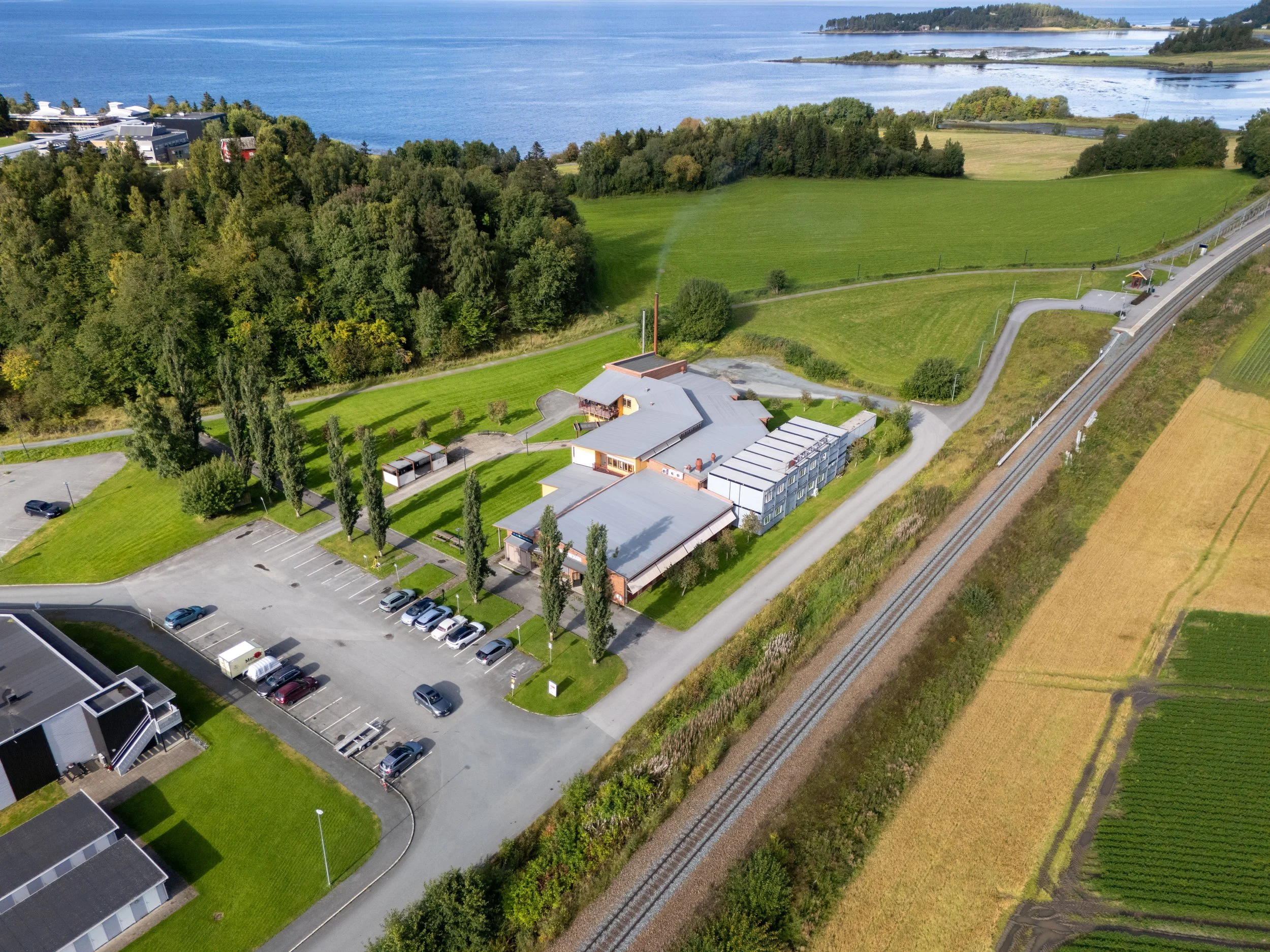 Aerial view of a rural area with a large body of water in the background, green fields, trees, and some buildings nearby. There are also train tracks visible on the right side of the image.
